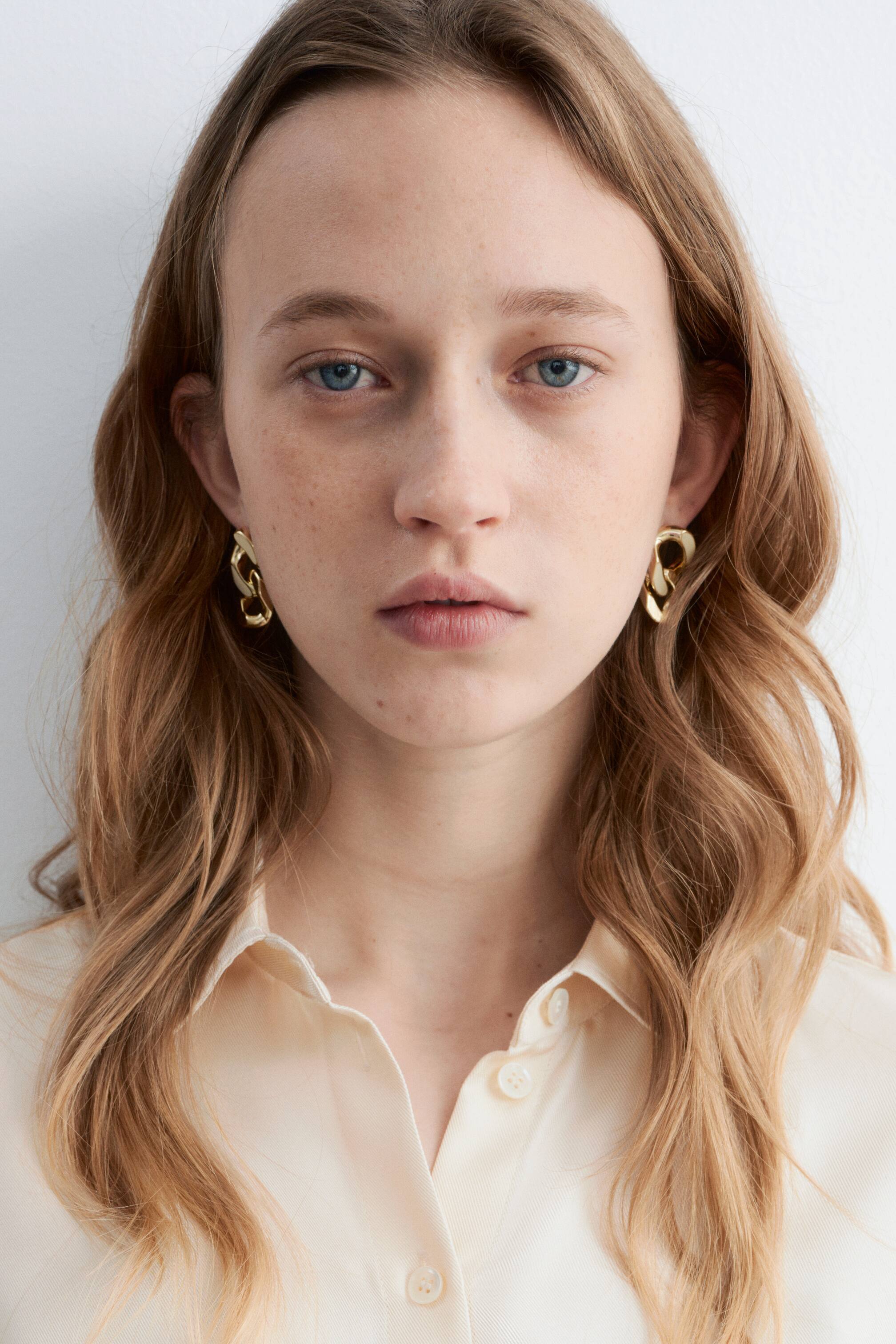 Grotere afbeelding bekijken: Goudkleurige abstracte schakel oorbellen hangen aan beide oorlellen, met een vloeiend, in elkaar grijpend ontwerp. Er wordt een licht crèmekleurig overhemd met kraag gedragen, waarvan de kraag zichtbaar is rond de hals.