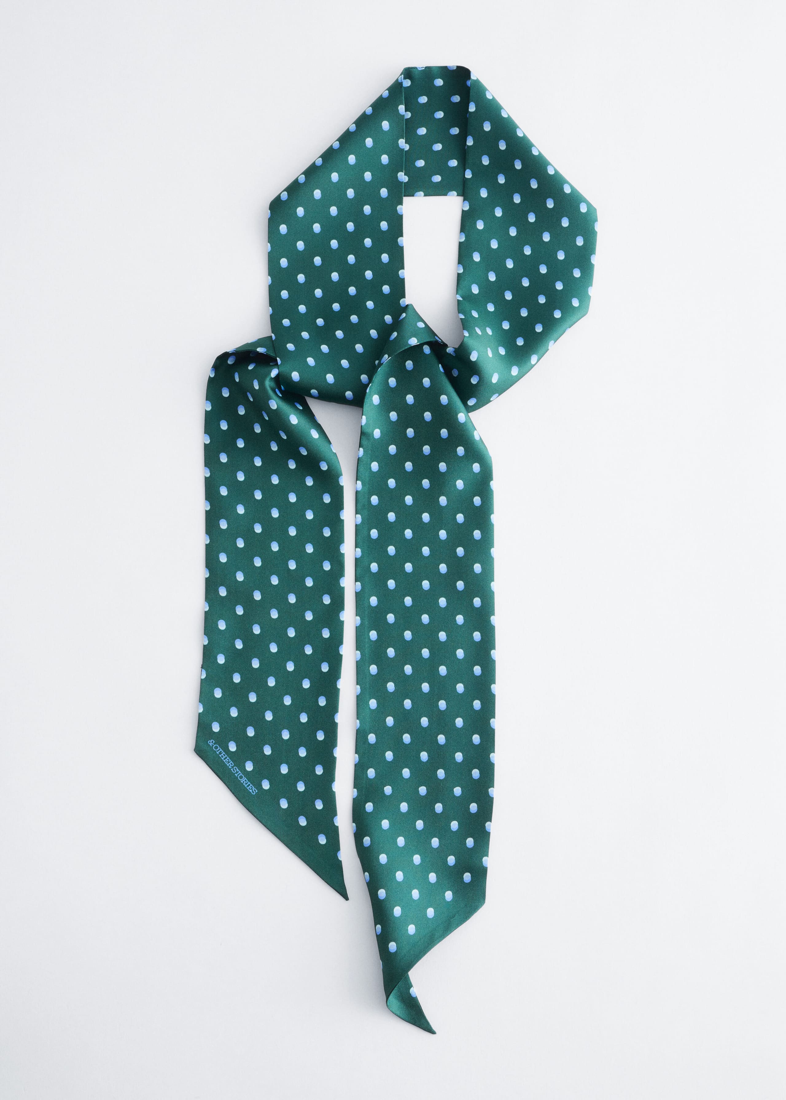 A slim green silk scarf featuring an all-over pattern of light blue polka dots is displayed in a loose knot, showcasing its long silhouette and pointed ends.