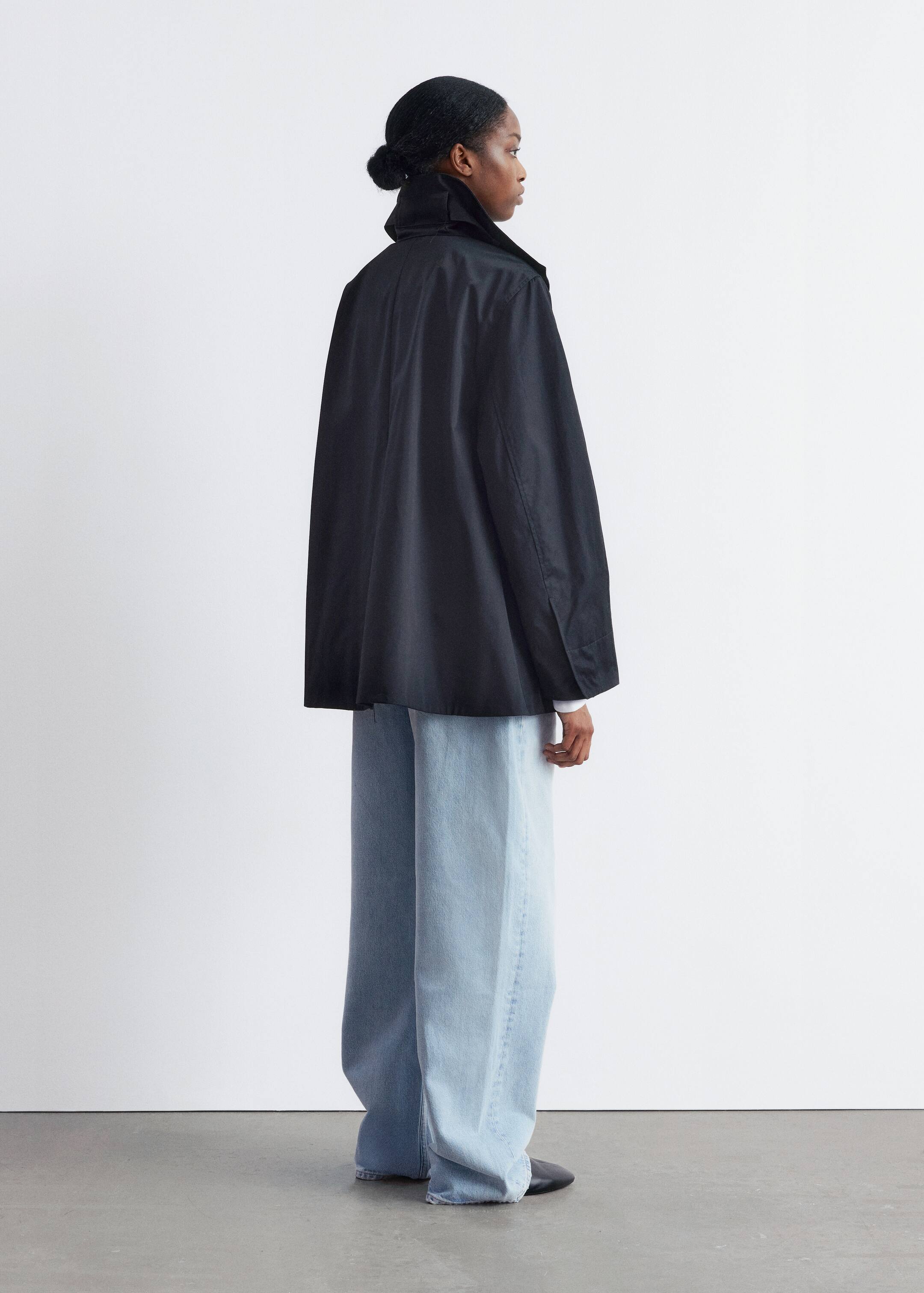 Grösseres Bild ansehen: Eine voluminöse schwarze Jacke mit einem hohen Stehkragen und weiten Ärmeln wird aus einem seitlich-hinteren Winkel präsentiert, wodurch ihr strukturierter, entspannter Fall hervorgehoben wird. Sie wird über hellblauen, weit geschnittenen Jeans getragen, die sich an den Knöcheln sammeln, wobei ein weißer Hemdmanschette unter dem Ärmel der Jacke hervorschaut.