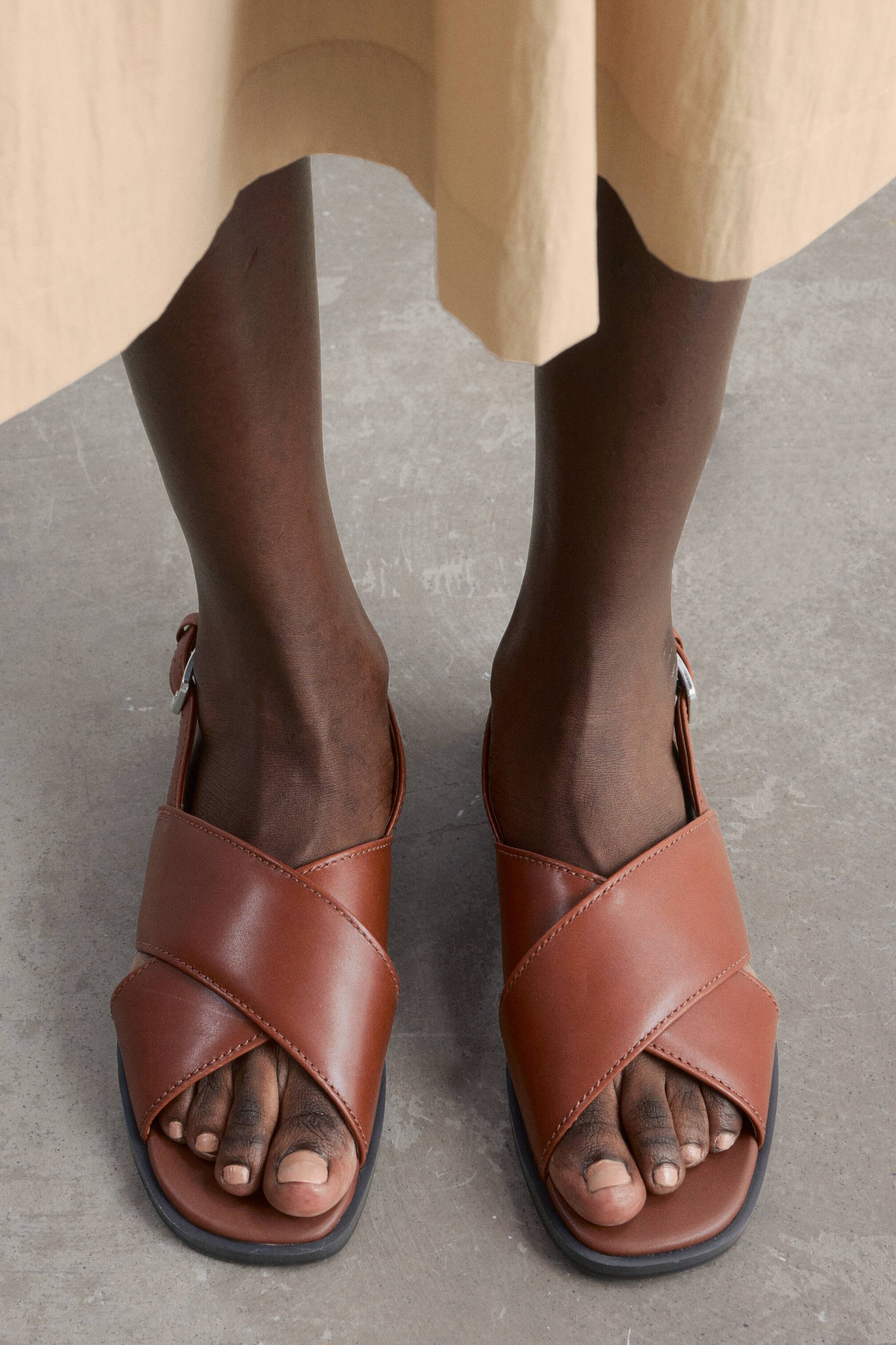 Προβολή μεγαλύτερης εικόνας: Δερμάτινα σανδάλια Crossover με καφέ δερμάτινα λουριά φοριούνται στα πόδια του μοντέλου. Τα λουριά διασταυρώνονται πάνω από το μπροστινό μέρος του ποδιού, ενώ ένα λουρί στον αστράγαλο με ασημένια αγκράφα ασφαλίζει κάθε σανδάλι, ορατό κάτω από το στρίφωμα ενός ανοιχτόχρωμου φορέματος.