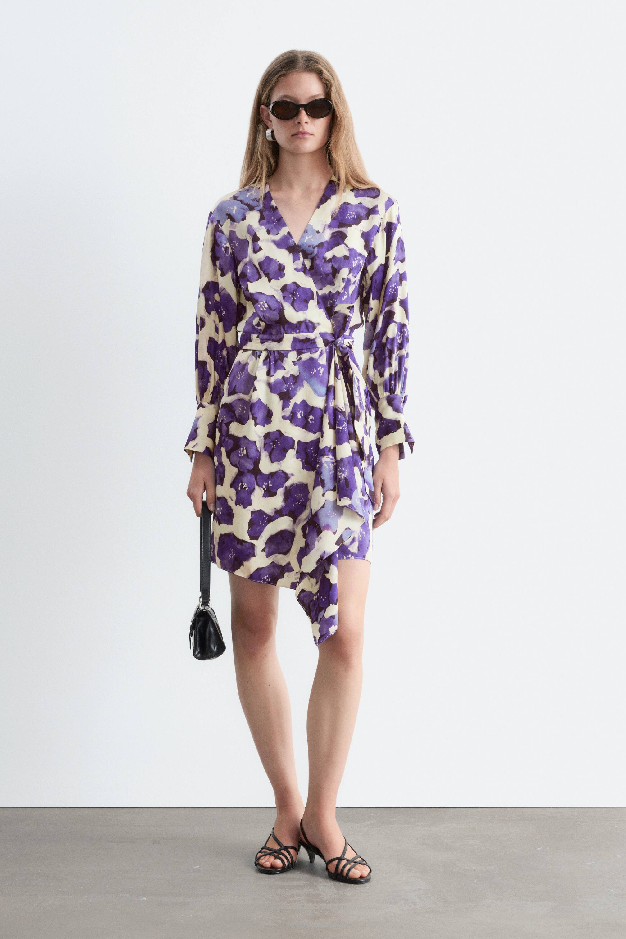 Agrandir l'image: Une robe portefeuille ornée d’un imprimé floral numérique violet et crème est présentée de face. Elle se distingue par un décolleté en V, des manches longues avec poignets froncés, ainsi qu’un devant croisé maintenu par une ceinture à nouer à la taille, et se termine par un ourlet asymétrique drapé.