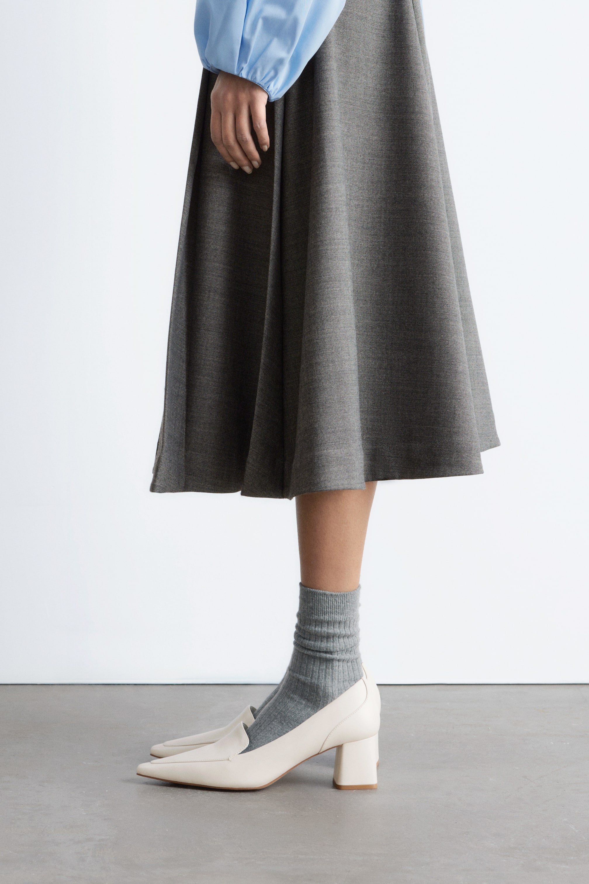 Größeres Bild ansehen: Ein melierter grauer A-Linien-Rock wird von dem Model getragen und schwingt sanft bis zu einer Midilänge. Er wird mit einem hellblauen langärmeligen Oberteil, grauen gerippten Socken und cremefarbenen Blockabsatz-Loafern kombiniert.