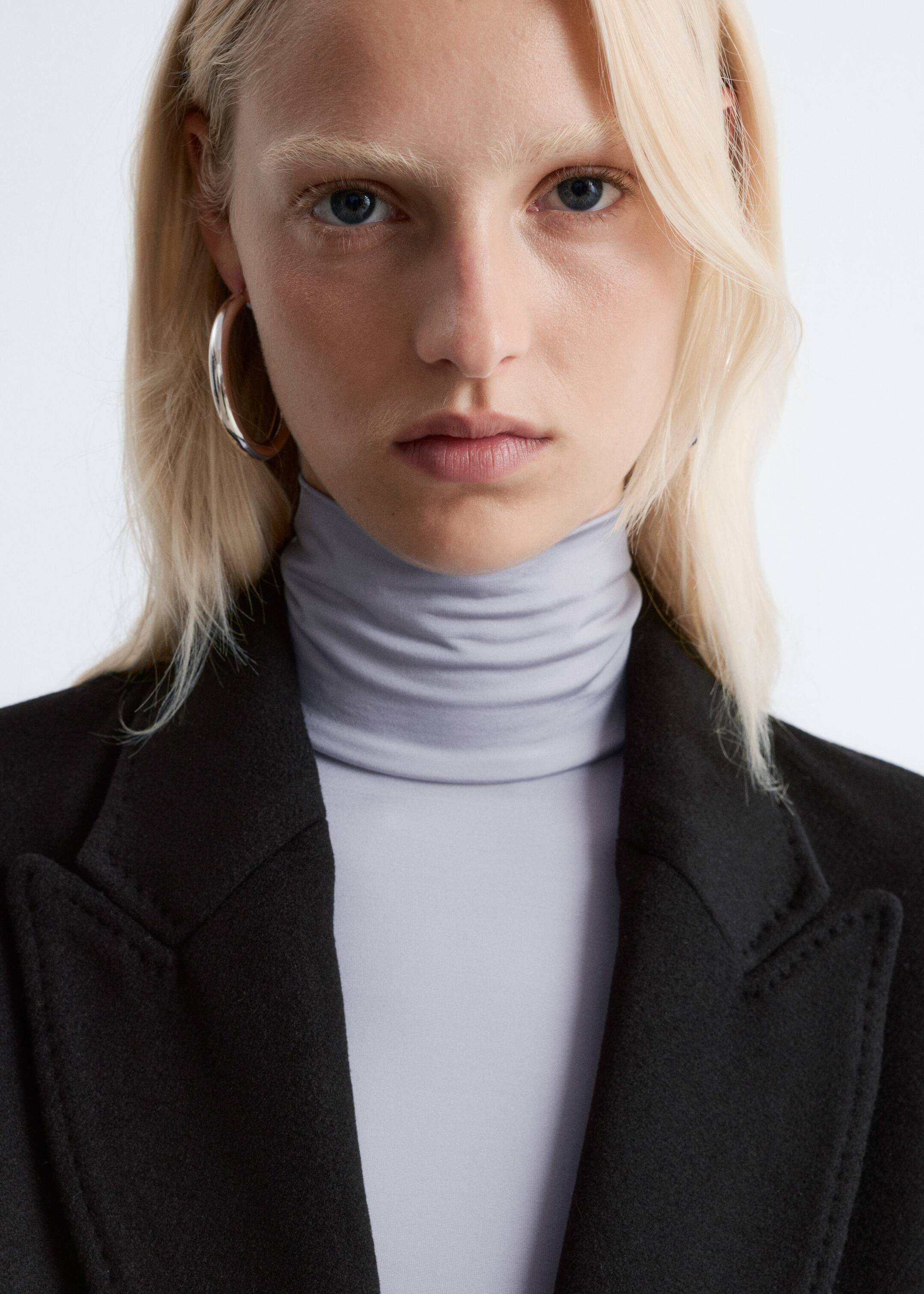 Agrandir l'image: Un manteau noir avec des coutures visibles sur ses revers crantés est porté par-dessus un col roulé gris clair, dont le col est délicatement plié.