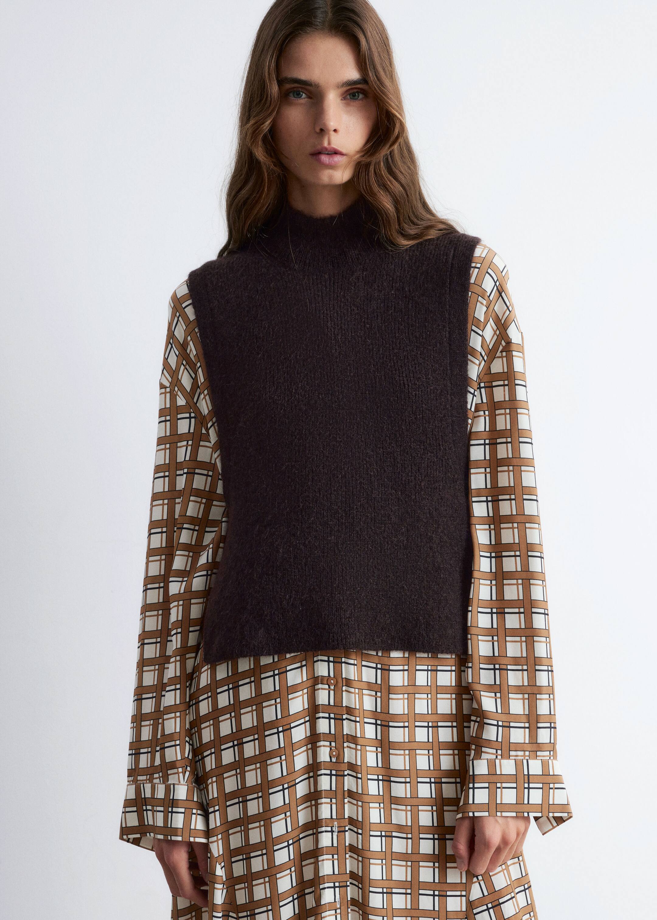 Größeres Bild anzeigen: Ein dunkelbraunes, gestricktes Mock-Neck-Weste wird von vorne präsentiert, über einem langärmeligen Kleid geschichtet. Die ärmellose Weste verfügt über ein strukturiertes Strickmuster und wirkt leicht verkürzt, während das darunterliegende Kleid mit einem braunen, weißen und schwarzen geometrischen Gittermuster versehen ist.