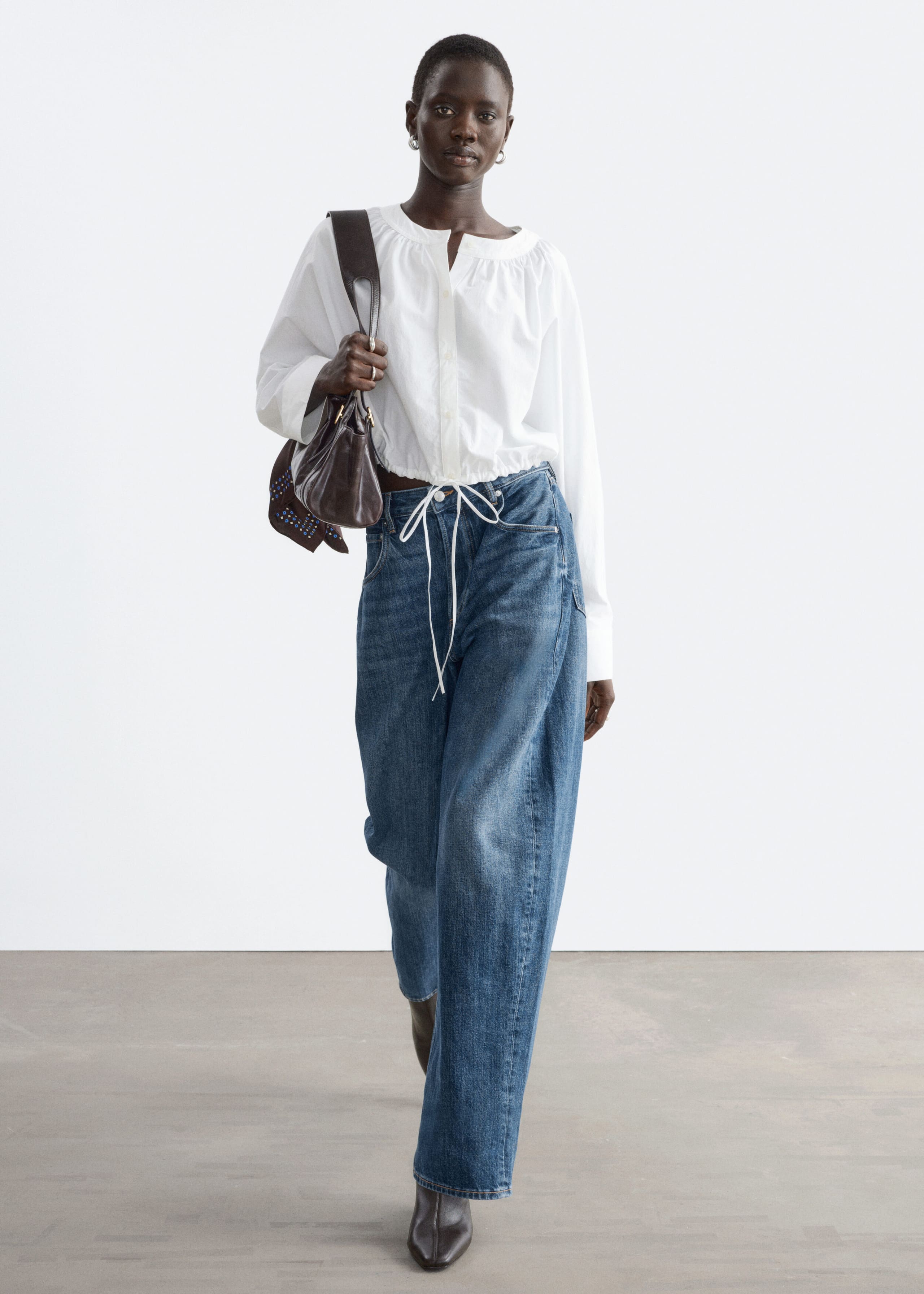 A white cropped blouse featuring a gathered neckline, a buttoned front placket and a tied drawstring hem is being worn by the model. The blouse is styled with wide-leg blue jeans, and the model is carrying a dark brown shoulder bag.