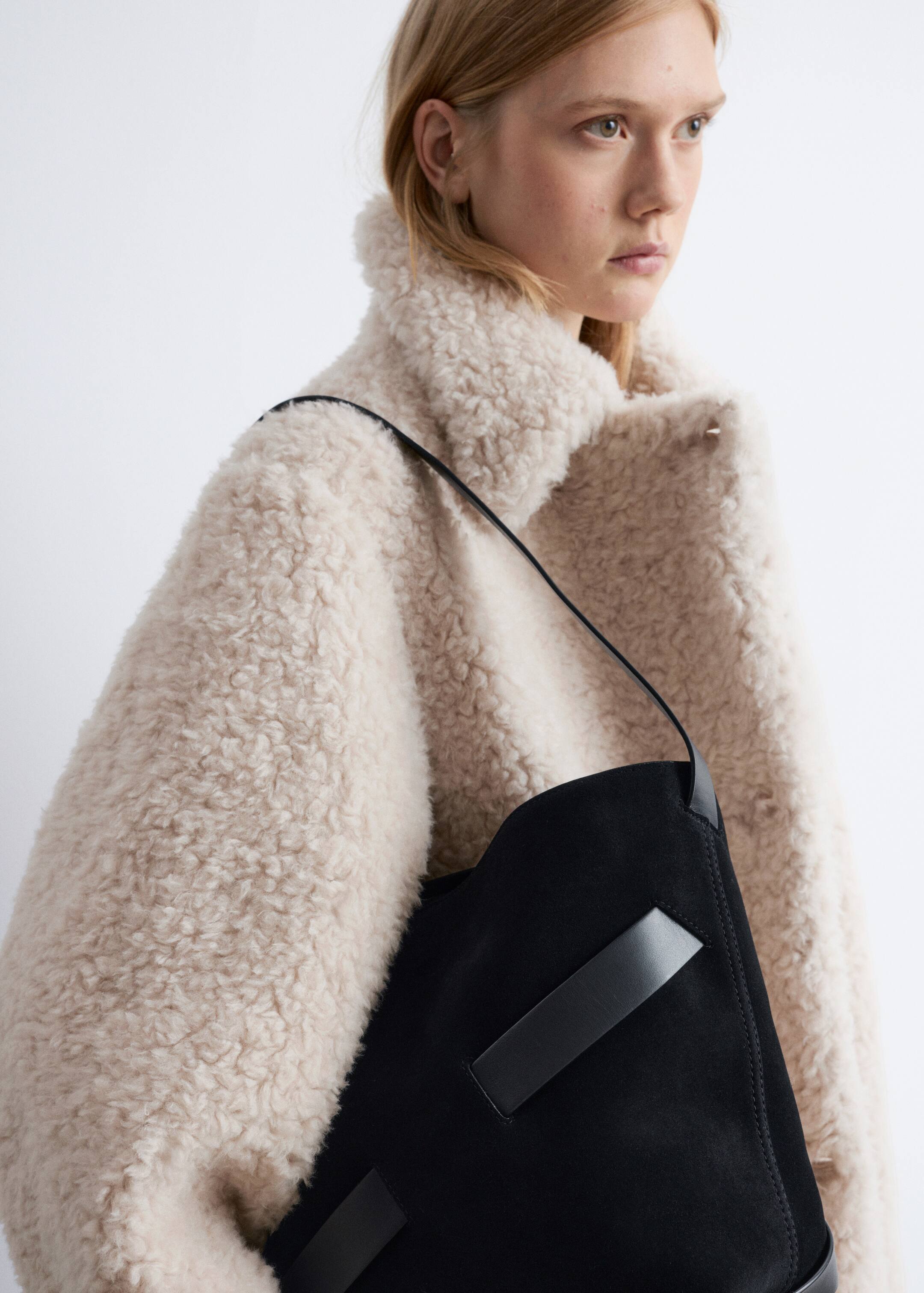 Grotere afbeelding bekijken: Een crèmekleurige jas in shearling-stijl wordt gepresenteerd vanuit een hoek aan de voorkant, waarbij de dikke, krullende textuur en brede kraag worden getoond. Een zwarte suède tas met een dunne riem en leren accenten rust tegen de voorkant van de jas.