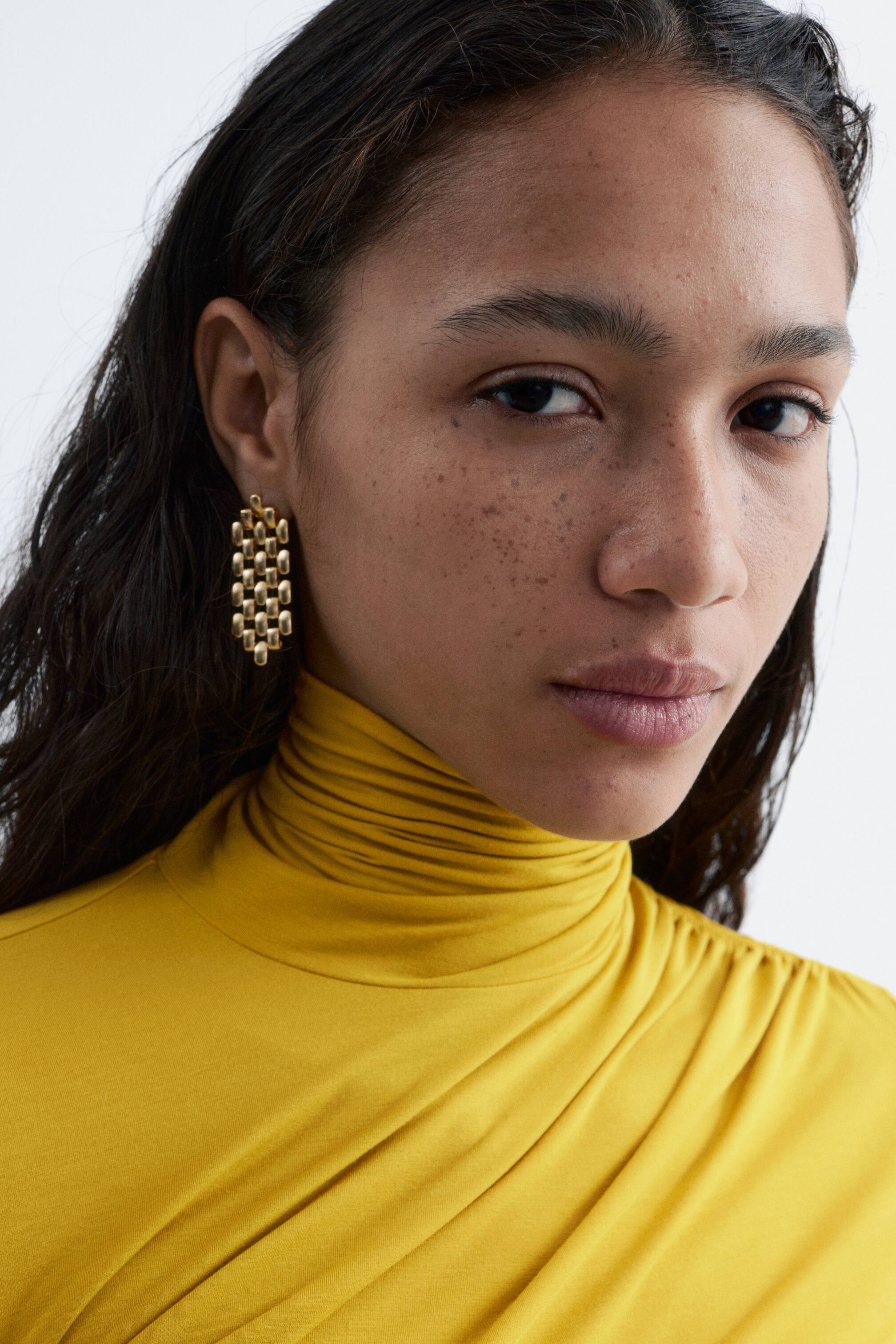 Agrandir l'image: Une boucle d'oreille pendante rectangulaire dorée est suspendue à l'oreille, sa surface présentant un motif complexe de maillons arrondis et entrelacés. Un haut à manches longues jaune moutarde est porté, avec un col roulé froncé et des fronces stratégiques sur l'épaule et la poitrine.
