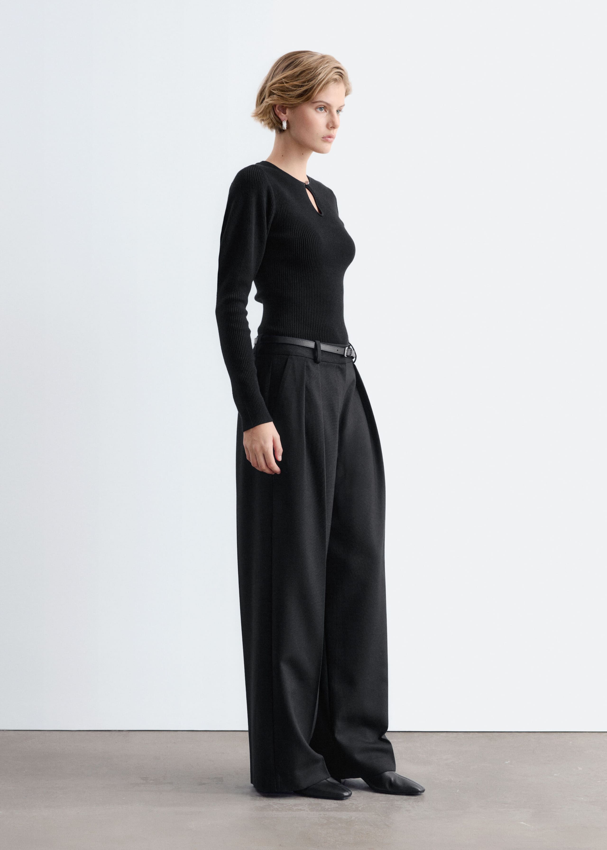 A black rib-knit top featuring a front keyhole opening and softly puffed shoulders is worn by the model. The close-fitting, long-sleeve top is tucked into wide-leg black trousers.