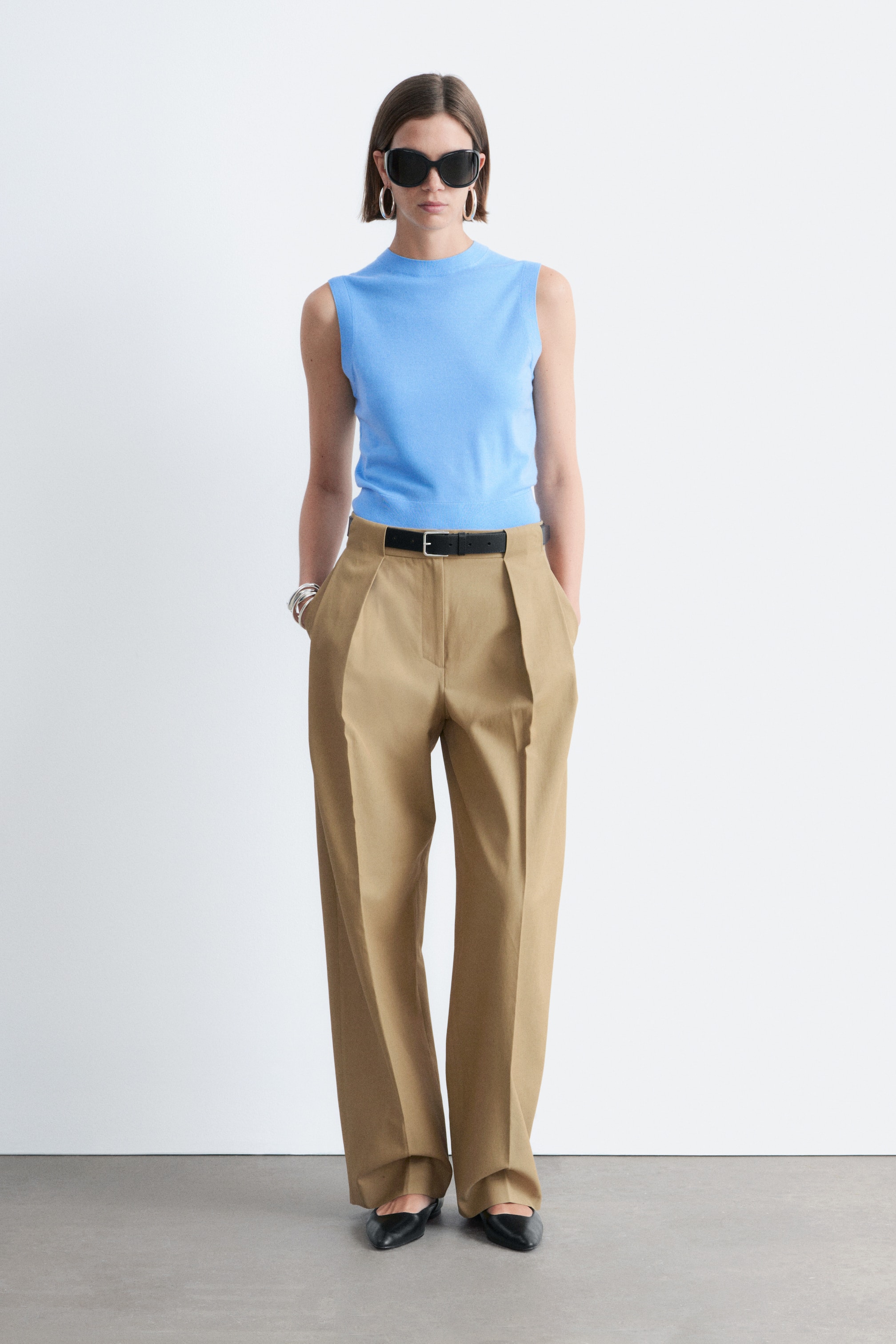Visualizza immagine più grande: Un top senza maniche a collo alto di colore azzurro è indossato infilato in pantaloni a vita alta color cachi, con doppia piega e gamba larga, stretti in vita da una cintura nera.