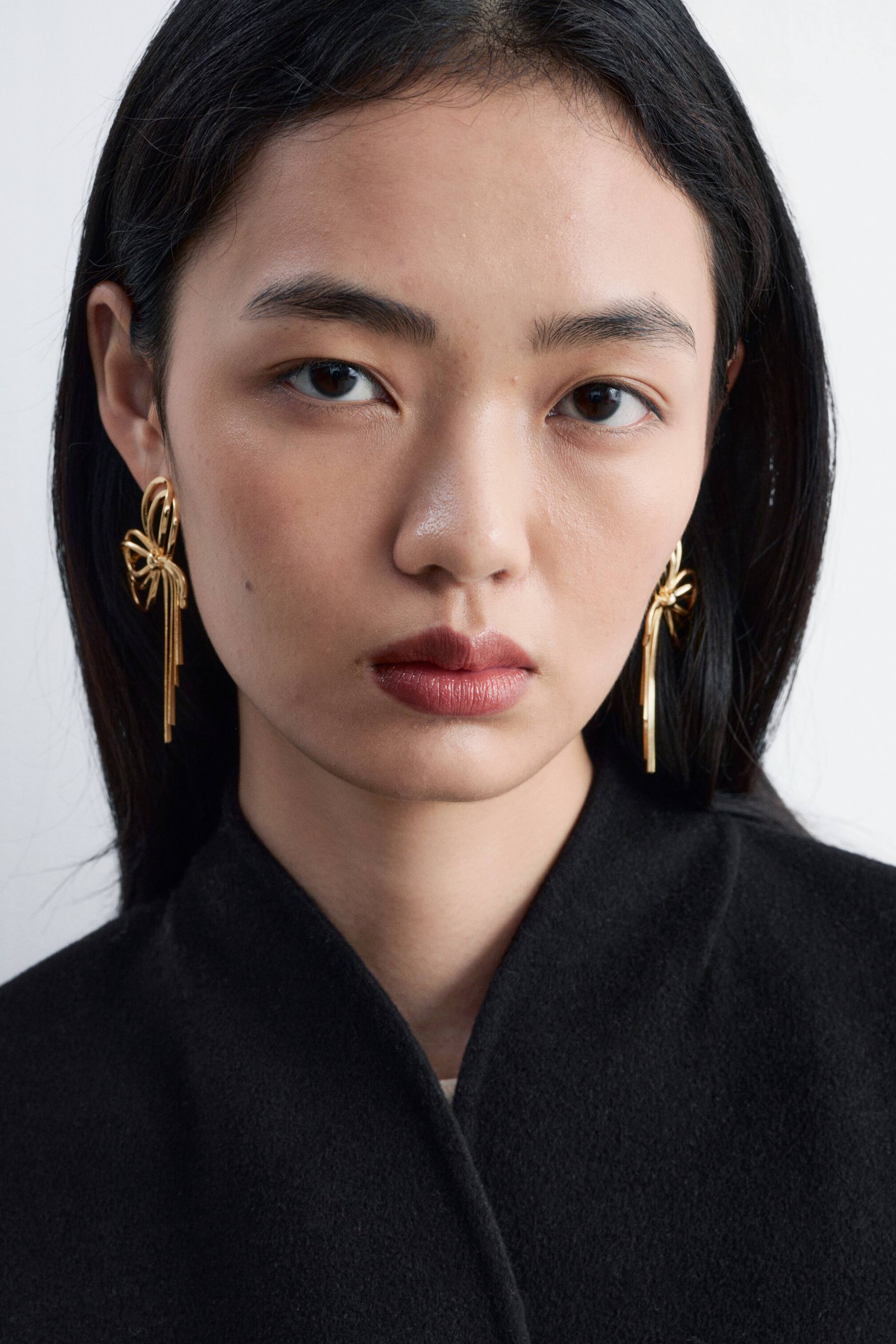 View larger image: Polished brass earrings shaped like bows, with long dangling metallic strands, are being worn by the model. The gold-toned earrings are highlighted by the model’s hair being tucked behind her ears and contrast against a high-necked black garment.