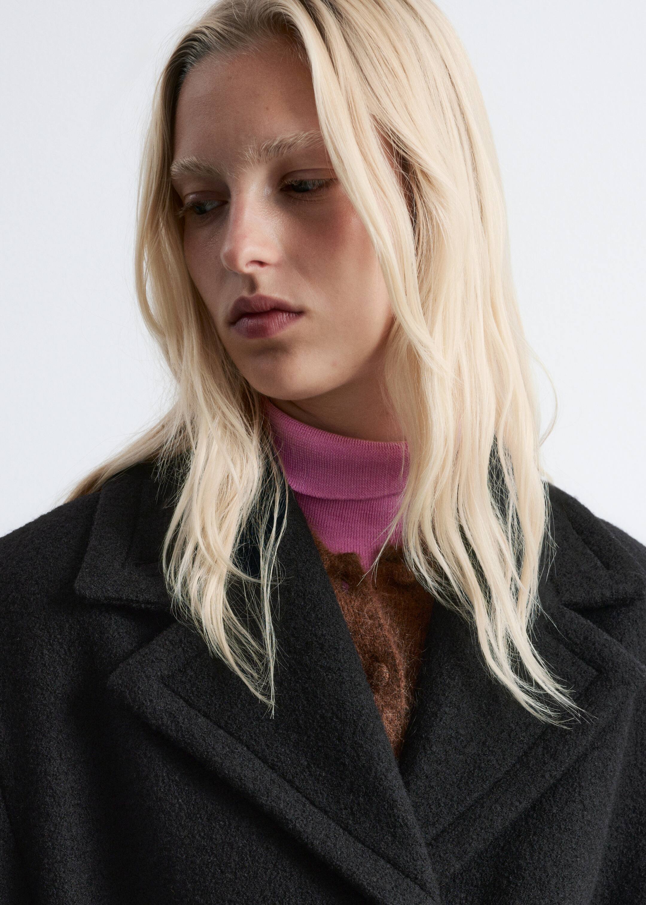 Agrandir l'image: Un manteau noir avec de larges revers crantés et un tissu texturé est présenté sous un angle avant-droit, porté ouvert sur un col roulé côtelé rose vif et un cardigan marron duveteux.