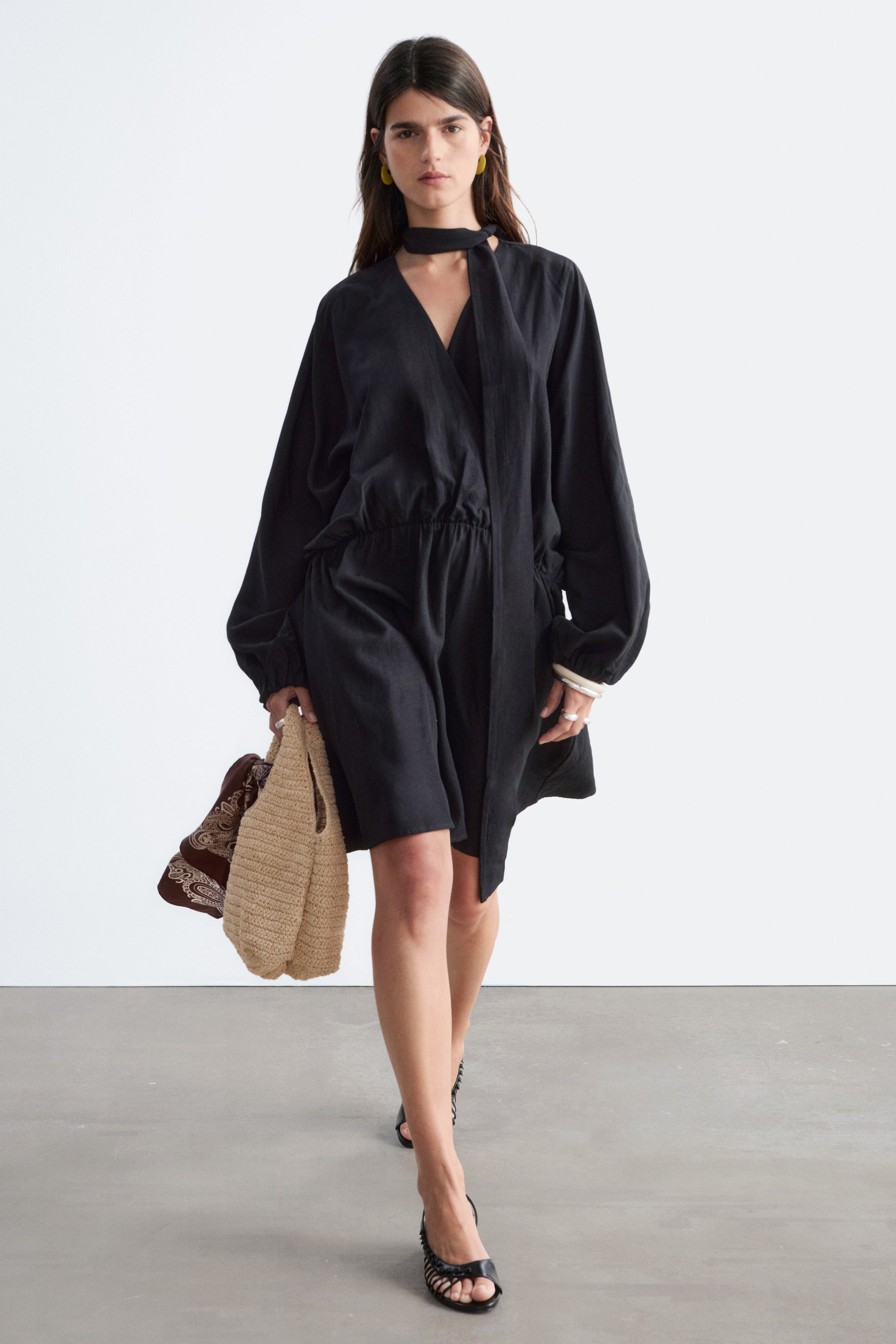 Agrandir l'image: Une robe légère noire présente un décolleté en V avec un long nœud intégré, des manches longues volumineuses avec des poignets froncés, et une taille élastiquée créant une silhouette cintrée.