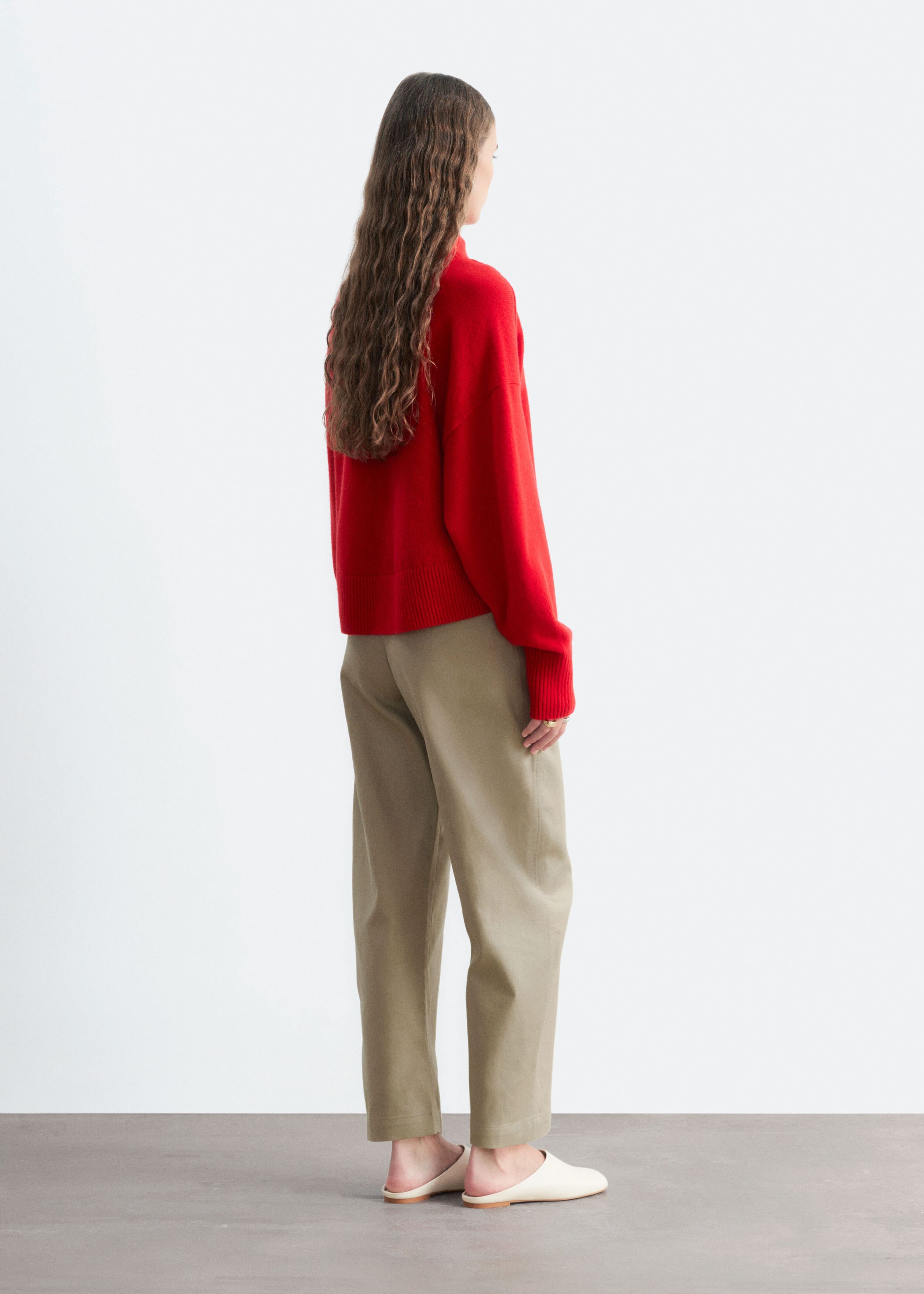 A red mock neck sweater with a relaxed fit and ribbed trims is being worn by the model, who is standing and facing away from the camera to show the back and side profile of the garment. The knitwear is styled with cropped khaki trousers and cream-colored mules.