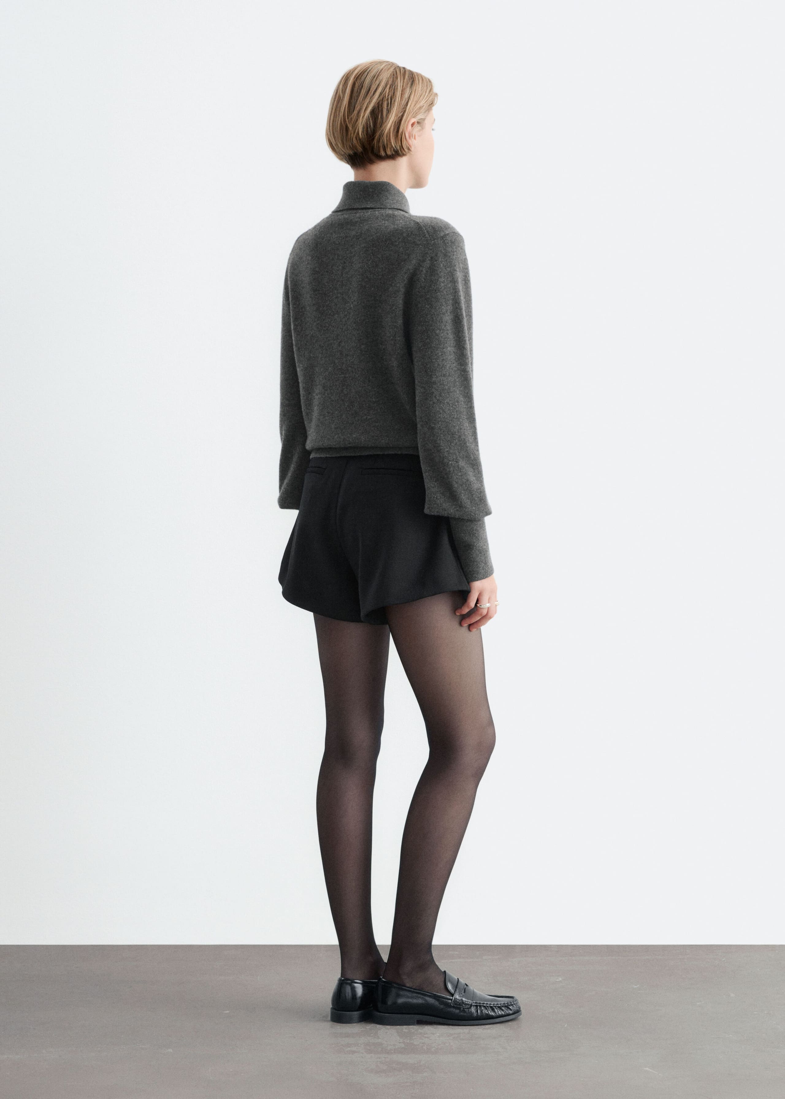 These black high-waisted shorts, featuring a soft A-line silhouette and a back welt pocket, are being worn by the model. The shorts are styled with a grey long-sleeved polo neck sweater, sheer black tights, and black leather loafers.