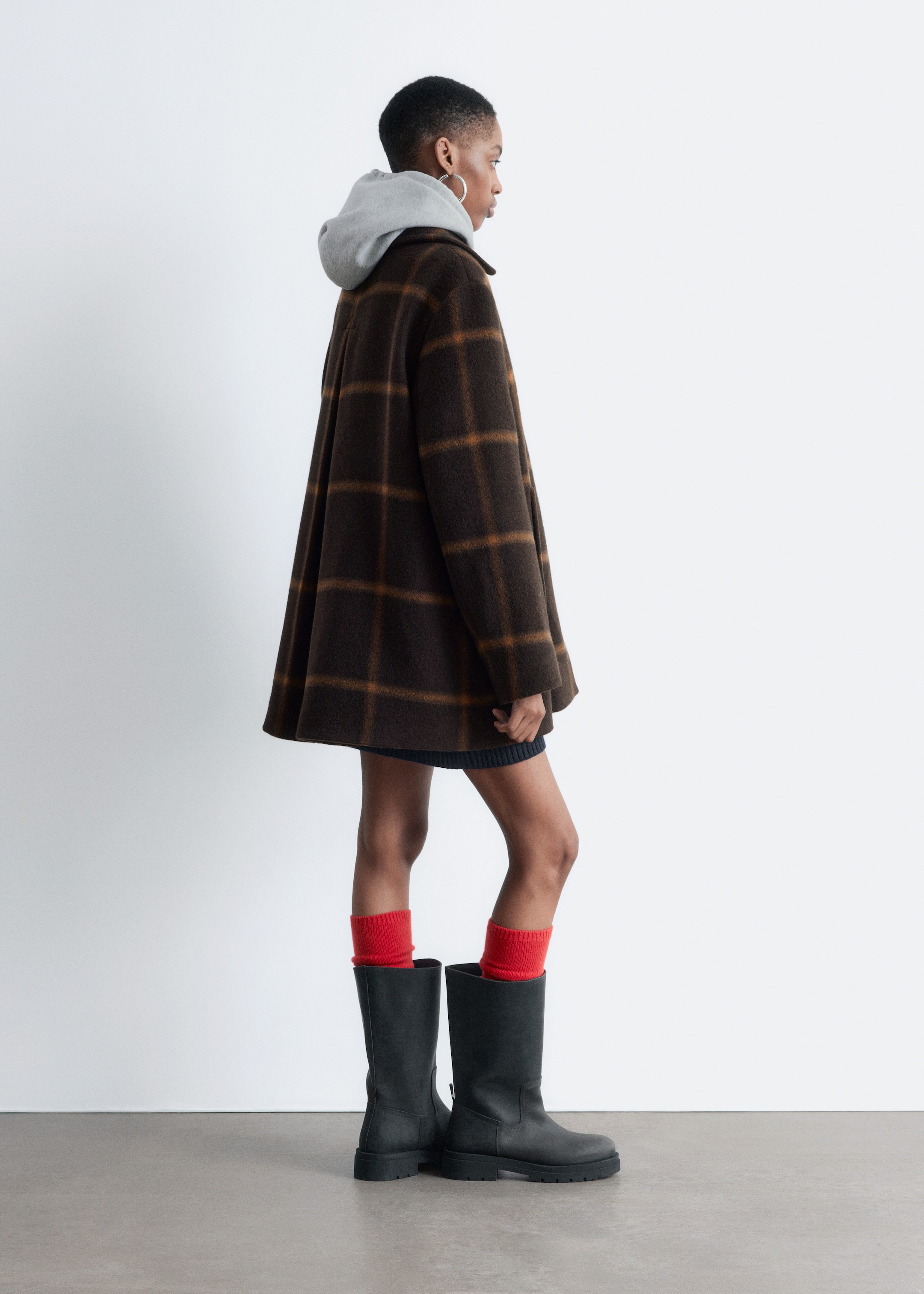 Grotere afbeelding bekijken: Een bruine en oranje geruite jas wordt gepresenteerd vanuit een zijaanzicht. Het heeft een oversized A-lijn silhouet met een rasterpatroon en valt boven de knieën. Een grijze hoodie wordt onder de jas gedragen, met de capuchon zichtbaar.