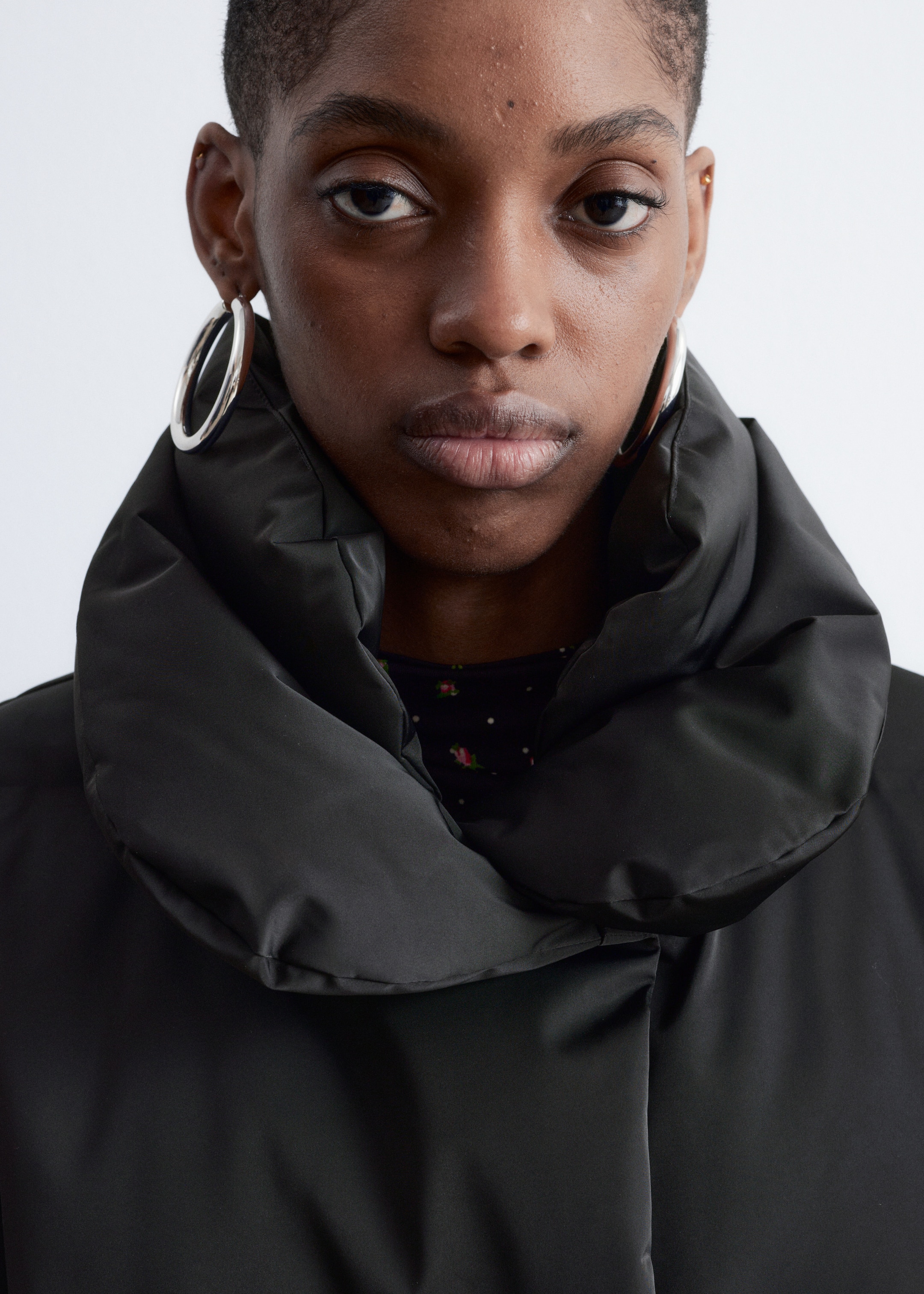 View larger image: A black padded jacket is featuring a high, voluminous stand-up collar and a smooth, matte fabric finish, seen from a front-facing perspective. It is being worn over a dark, patterned top with small floral and dot motifs, which is partially visible at the neckline.
