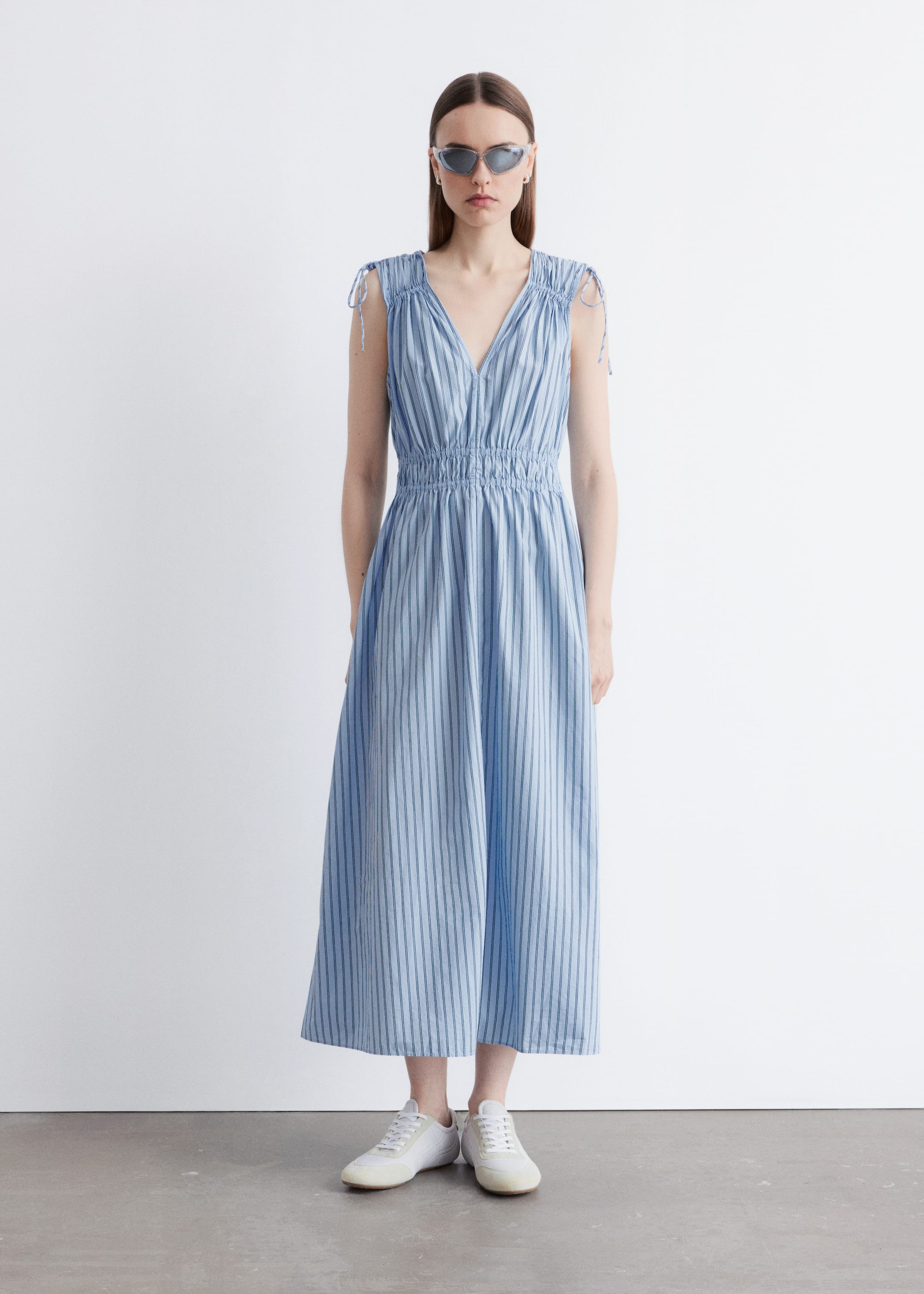 Größeres Bild ansehen: Ein hellblaues Midikleid mit vertikalen dunkelblauen Streifen wird frontal präsentiert. Es verfügt über einen V-Ausschnitt, Bindebänder an den Schultern und ein gerafftes Oberteil, das in eine elastische, gesmokte Taille übergeht. Der A-Linien-Rock fließt in Midilänge und endet über weißen Sneakers.