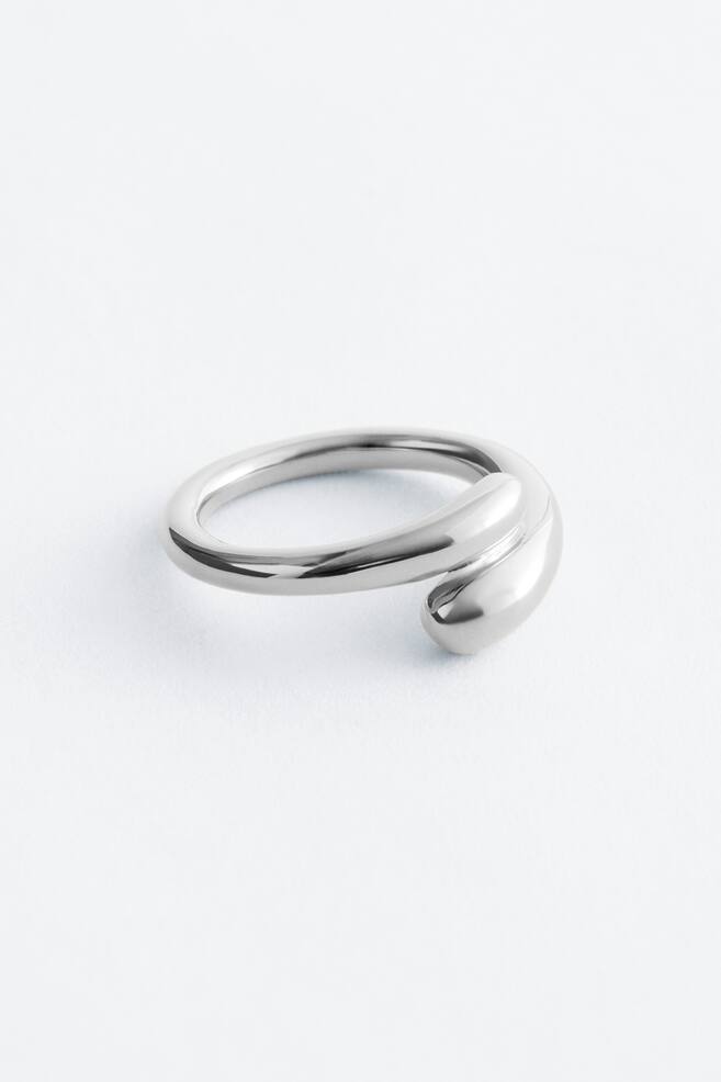 Een zilverkleurige ring toont een open ontwerp, met een slanke, gepolijste band die zich buigt naar een grotere, afgeronde, druppelvormige element dat de band overlapt. - 1