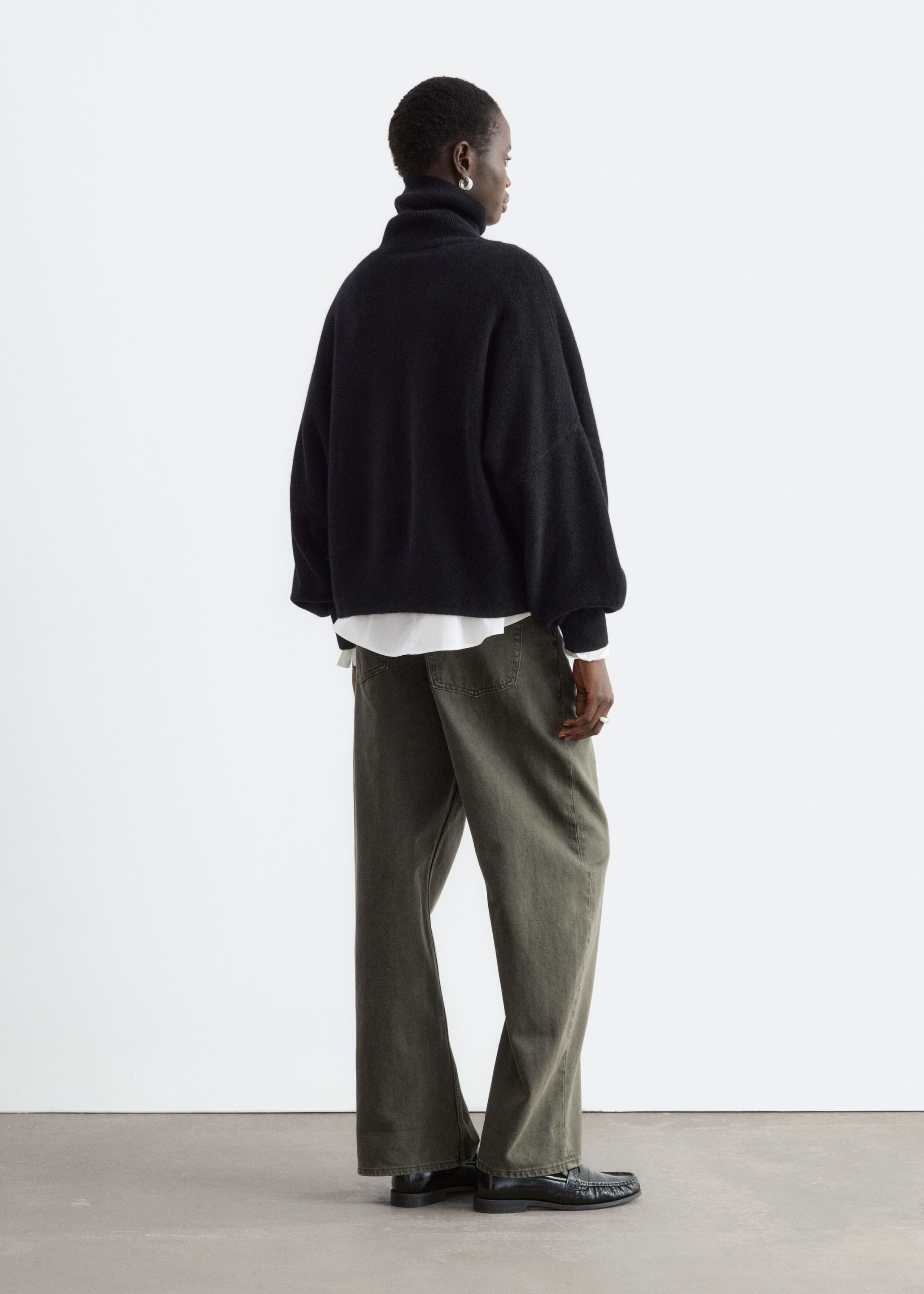The model is wearing a black oversized jumper with a folded rib turtleneck and dropped armholes, shown from a back three-quarter view. The jumper is layered over a white long-sleeved shirt with a visible hem and paired with wide-leg green trousers.