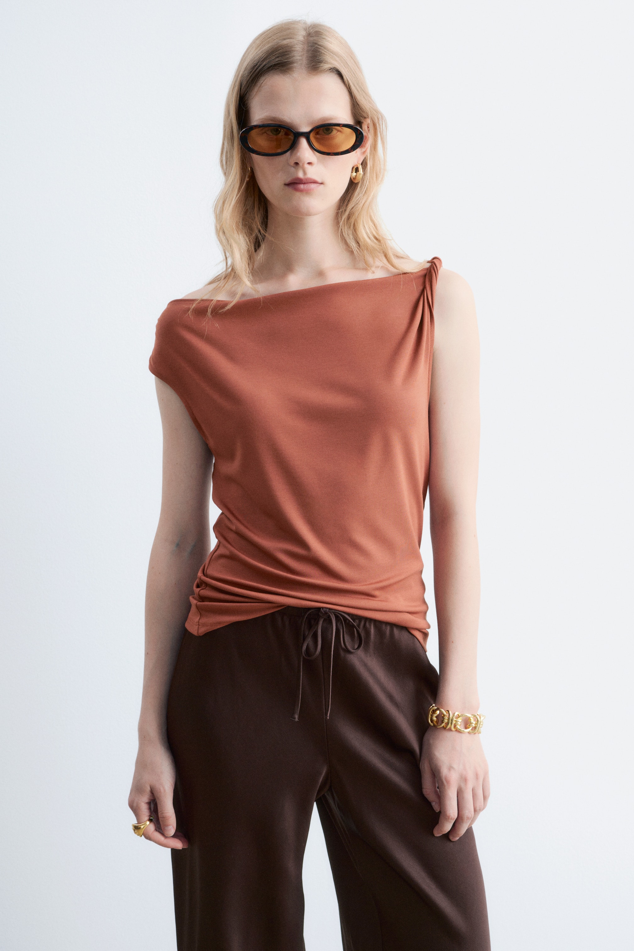 Agrandir l'image: Un haut couleur rouille présente un décolleté asymétrique avec une bretelle torsadée sur l'épaule droite et un style drapé tombant sur l'épaule gauche. Il met en avant une coupe douce et drapée avec une manche courte à revers sur le côté gauche.