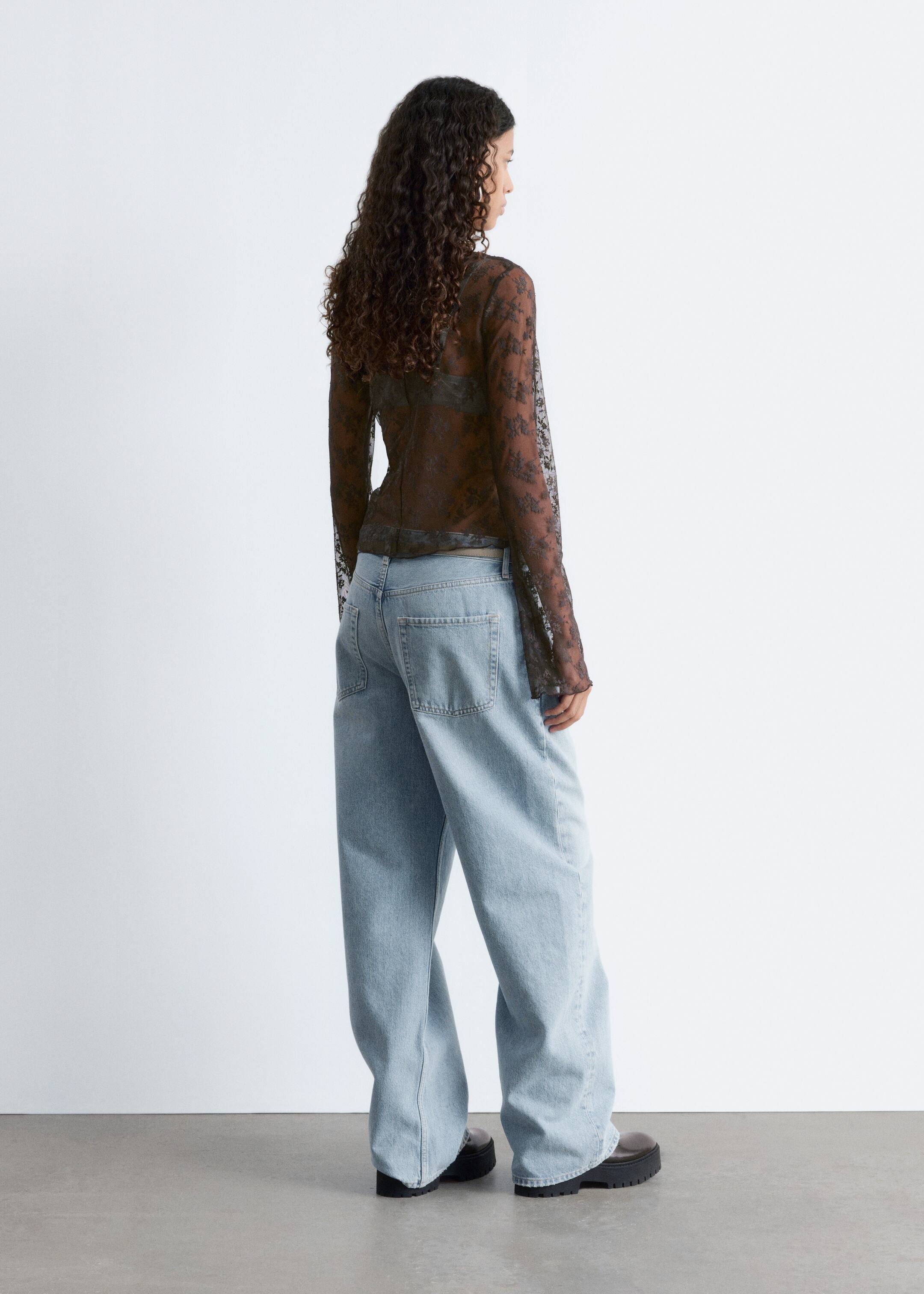 Agrandir l'image: Un haut en dentelle noire transparente est porté, présentant un motif complexe, des manches longues, et révélant un soutien-gorge sombre en dessous. Un jean à jambes larges délavé est porté, montrant des poches à l'arrière et une coupe décontractée, s'étendant pour couvrir le haut de chaussures sombres à semelles épaisses.