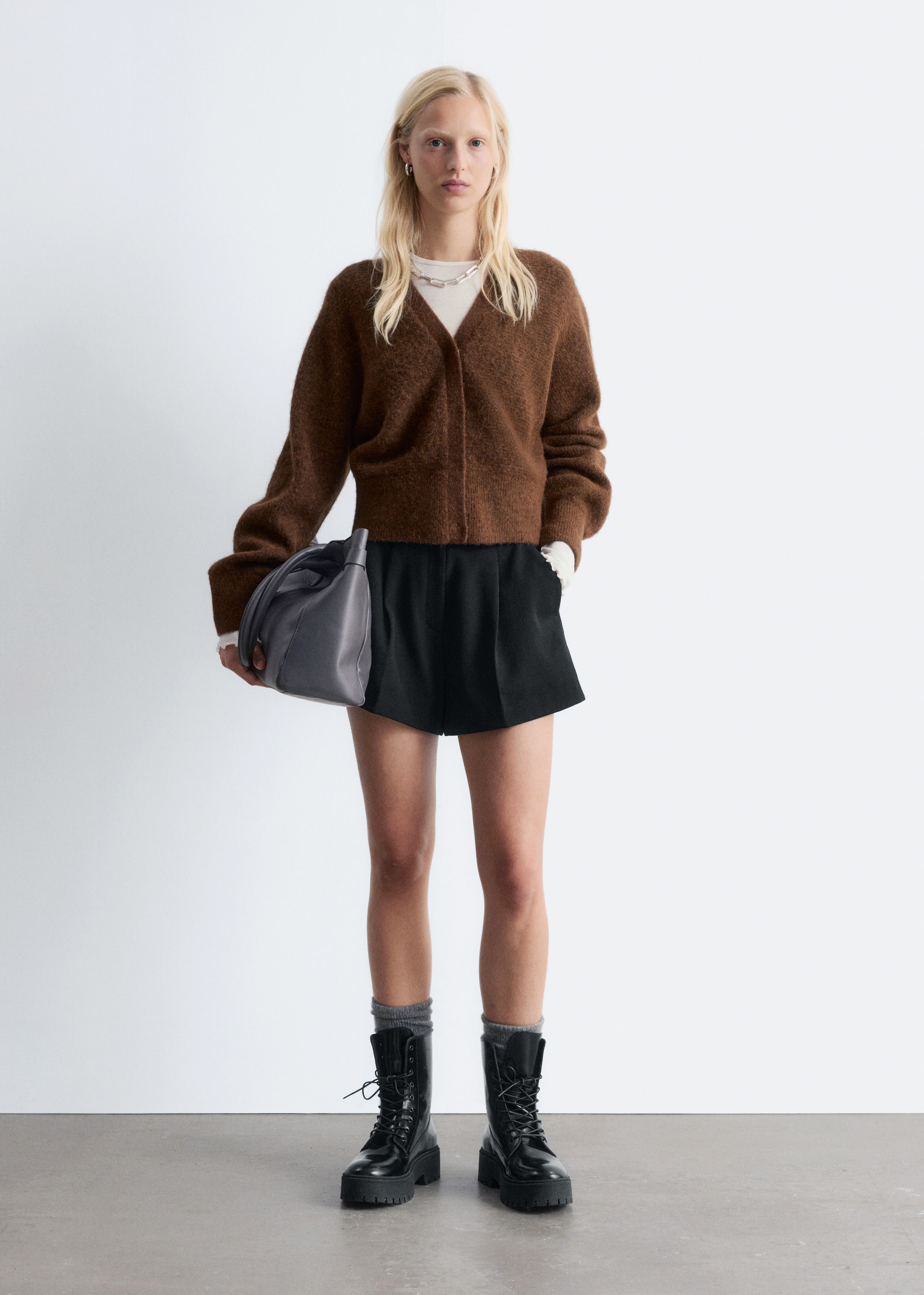 Ingrandisci l'immagine: L'outfit, presentato frontalmente, include un cardigan marrone a V peloso e corto, indossato sopra un top bianco a maniche lunghe con polsini visibili. È abbinato a pantaloncini neri a pieghe, stivaletti neri massicci con lacci, abbinati a calzini grigi e una borsa grigia a pieghe.