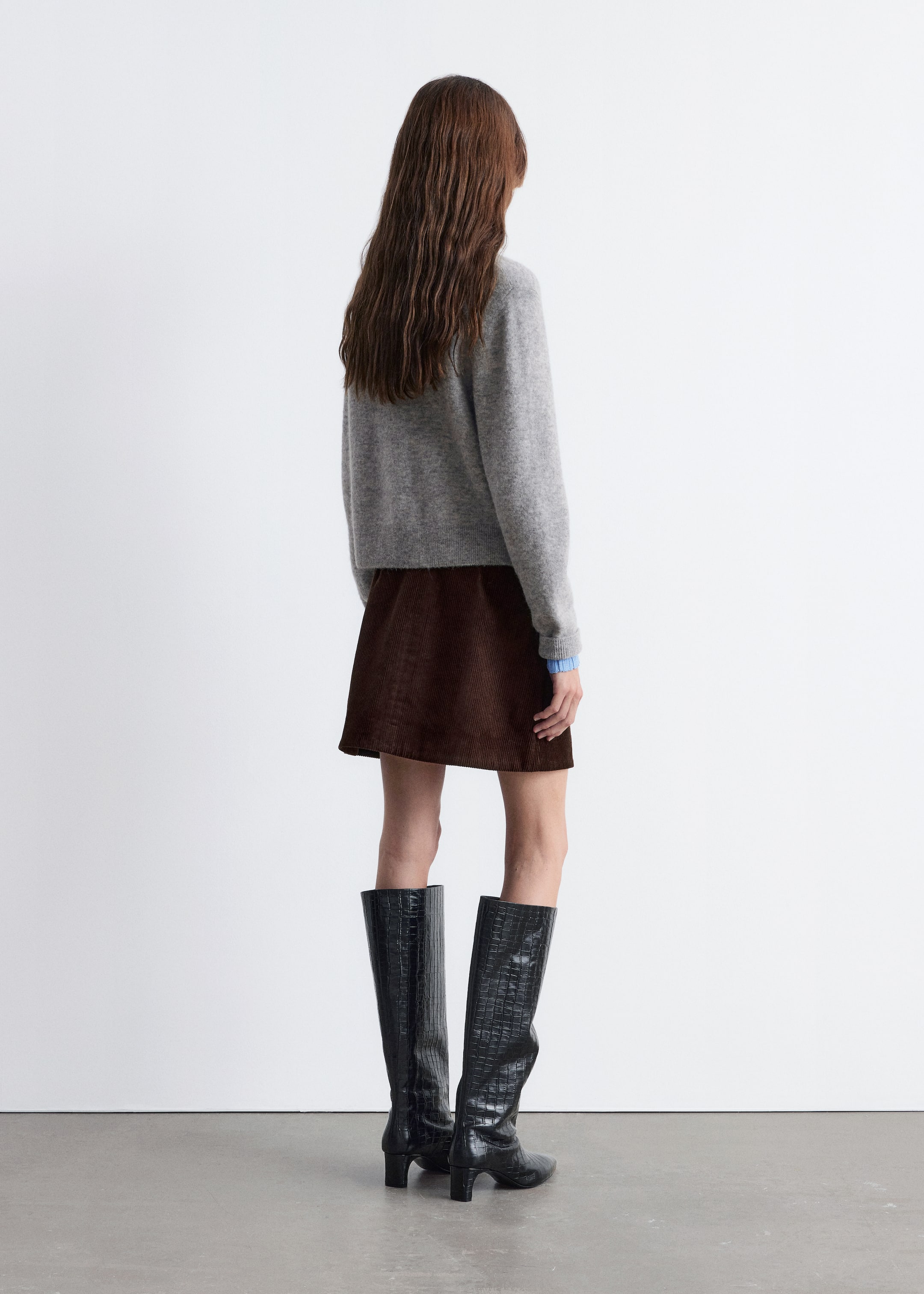 Agrandir l'image: Un pull gris clair, ample, avec un poignet bleu clair dépassant par-dessous, est montré de dos. Une mini-jupe trapèze en velours côtelé marron foncé est assortie à des bottes noires jusqu'aux genoux, qui présentent une texture embossée et des talons blocs.