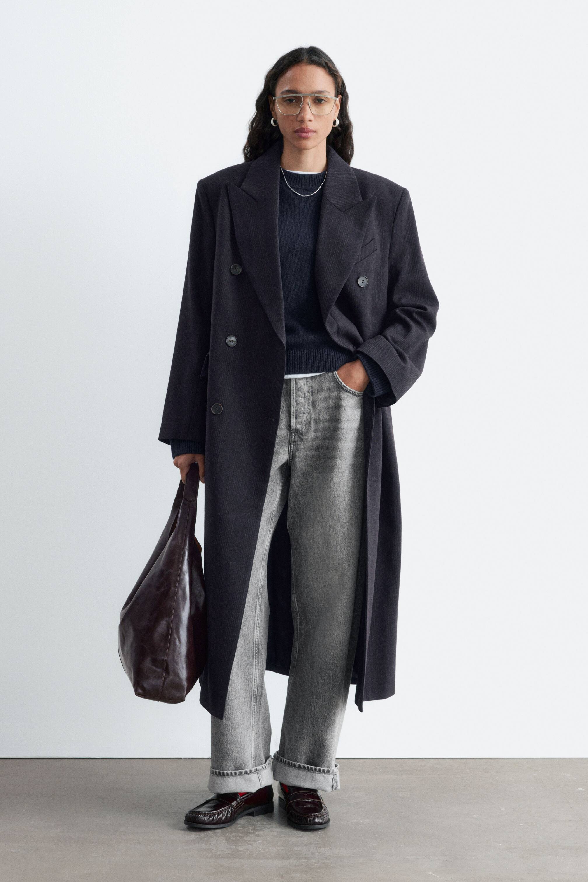 Se større billede: En mørkegrå, dobbeltknappet oversized frakke med markerede skuldre og spidse revers bæres åbent af modellen. Modellen har frakken som et lag udenpå en mørk sweater med rund hals og grå jeans og bærer samtidig en mørkebrun tote-taske i læder.
