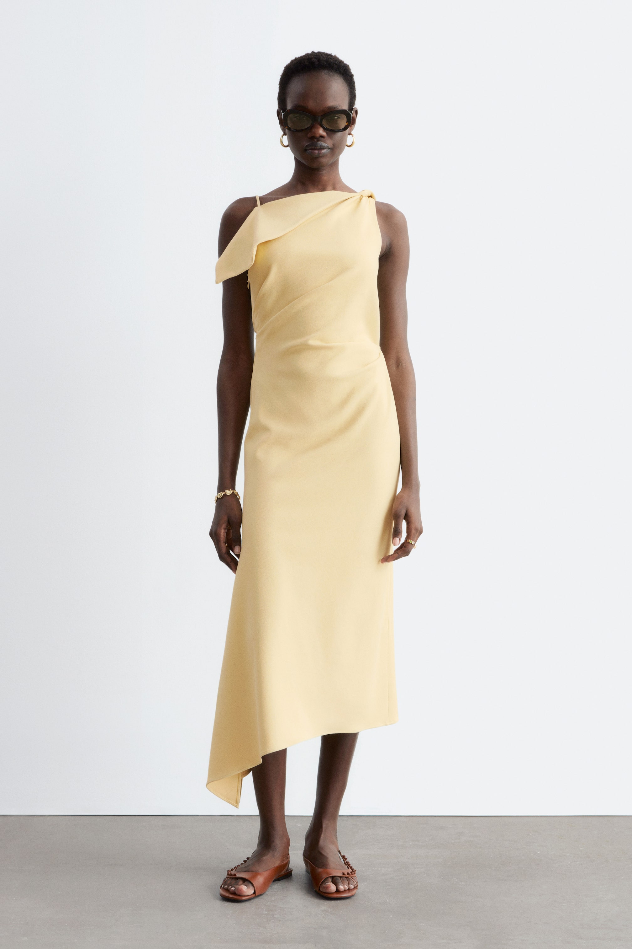 Grotere afbeelding bekijken: Een lichtgele midi-jurk wordt frontaal getoond, met een asymmetrische halslijn en een gedrapeerd detail op één schouder, diagonale drapering over het lijfje, en een asymmetrische midi-zoom.