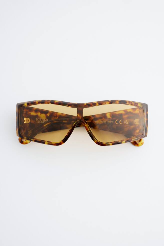 A pair of rectangular shield-style sunglasses is facing forwards, featuring a thick tortoiseshell-patterned frame in shades of brown and yellow. The sunglasses are showcasing light amber-tinted lenses with gold-toned lettering visible on the interior. - 1