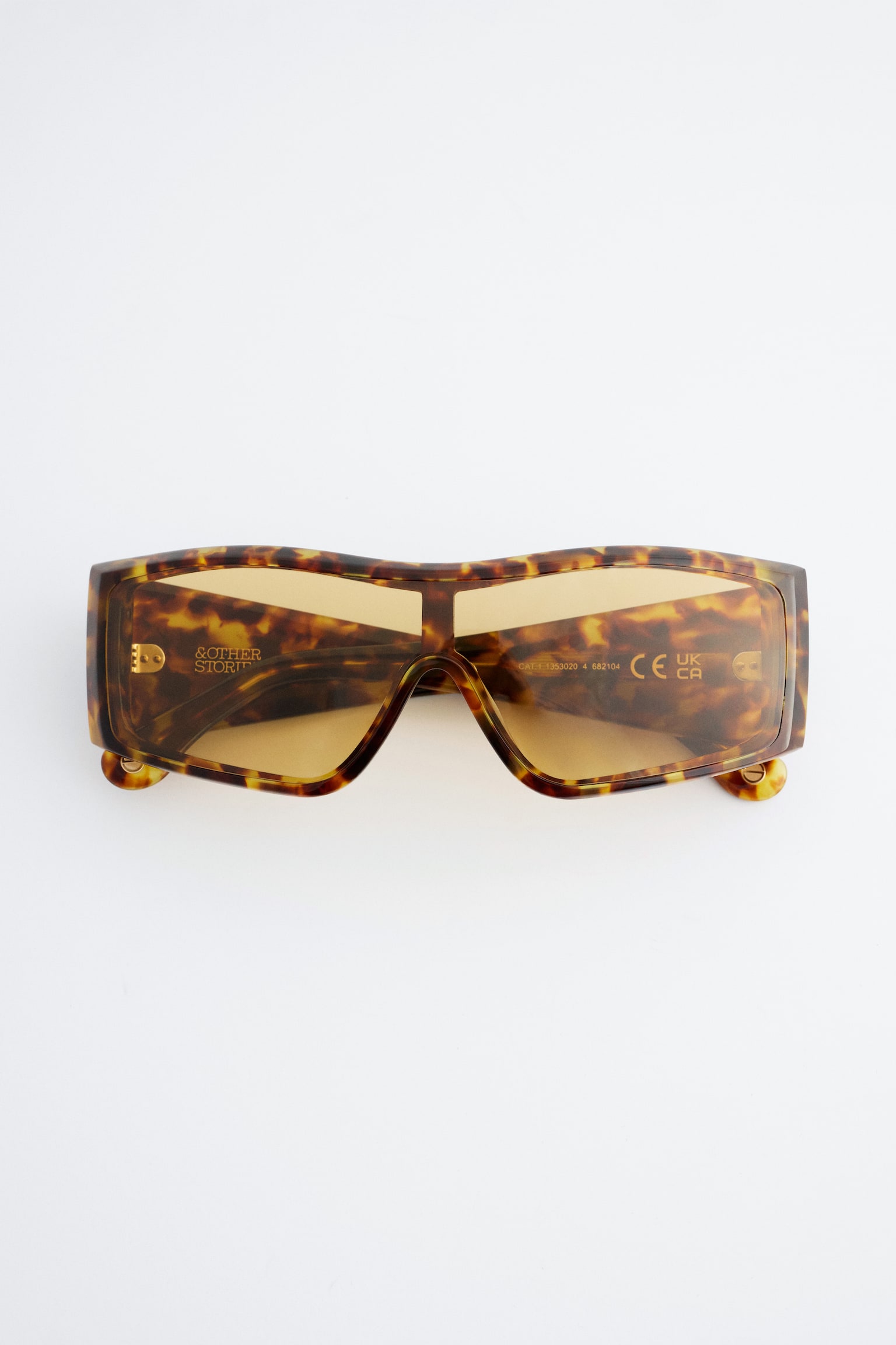 Une paire de lunettes de soleil rectangulaires de style écran est présentée de face, avec une monture épaisse à motif écaille de tortue dans des tons de brun et de jaune. Les lunettes de soleil mettent en valeur des verres légèrement teintés dâambre, avec des lettrages aux tons dorés visibles à lâintérieur.