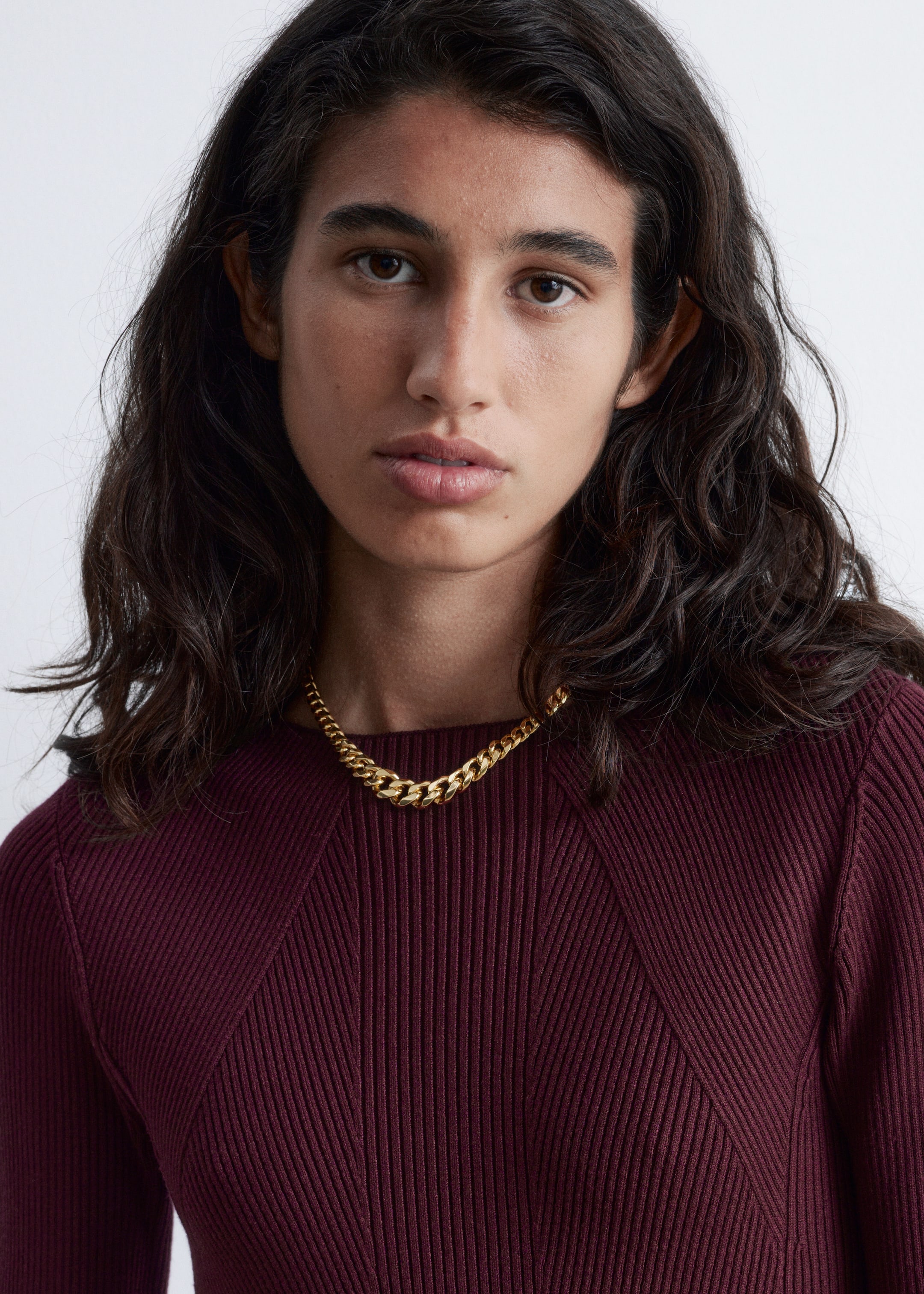 Se større billede: En bordeaux strikket mini-kjole med mock neck, der har en ribbet finish og diagonale sømlinjer på den øvre overdel, bæres af modellen. Et guldkædehalskæde er lagdelt over kjolen.