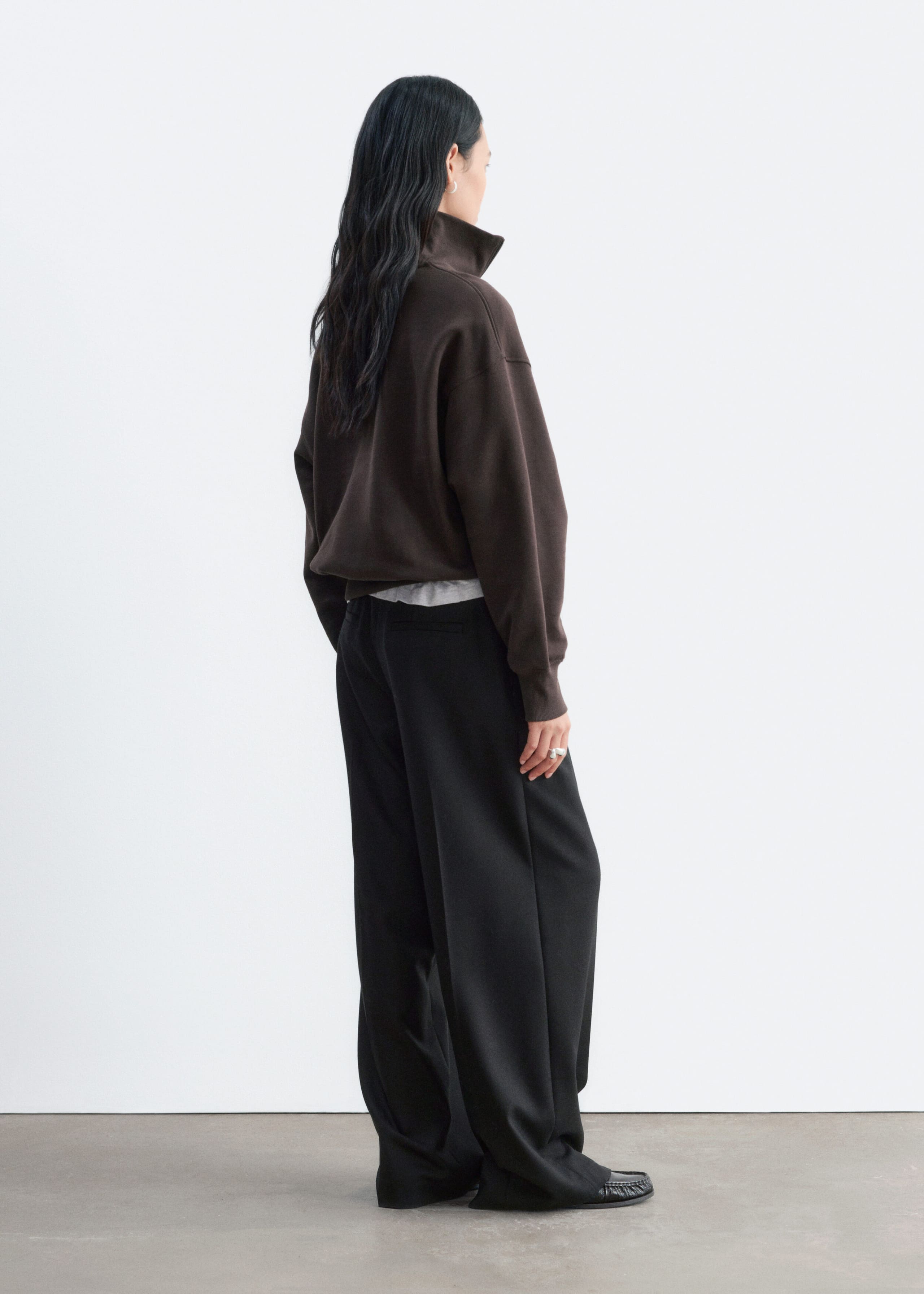 A dark brown, relaxed-fit sweatshirt is worn by the model, shown from a three-quarter rear angle. The sweatshirt features a high stand collar and ribbed cuffs, and is layered over a light grey top and paired with wide-leg black trousers.