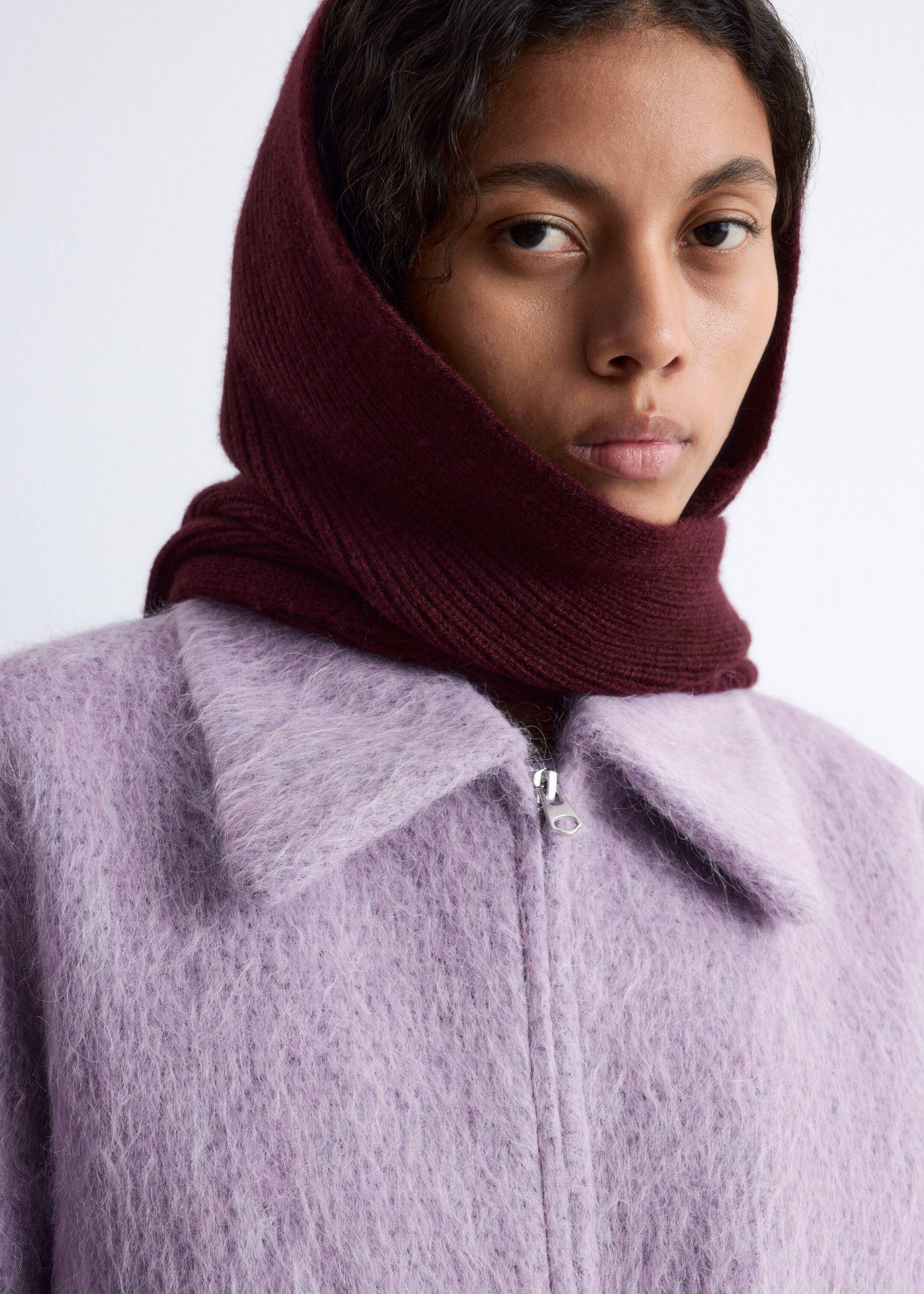 Visualizza immagine più grande: Una giacca lavanda, caratterizzata da una texture morbida e un ampio collo, è visibile dalla parte anteriore, con una zip argentata visibile al collo. Uno scaldacollo a maglia a coste color bordeaux copre la testa e il collo, sovrapponendosi al colletto della giacca.