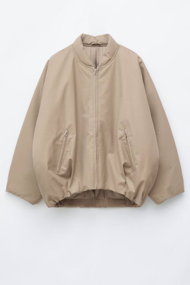 Een beige bomberjack is van voren te zien en toont een oversized, volumineus silhouet met een volledige ritssluiting aan de voorkant. Het jack heeft een gewatteerde opstaande kraag, verlaagde schouders met wijde mouwen, twee schuine ritszakken en een elastische zoom die een ronde, bolvormige look creÃ«ert. - 1