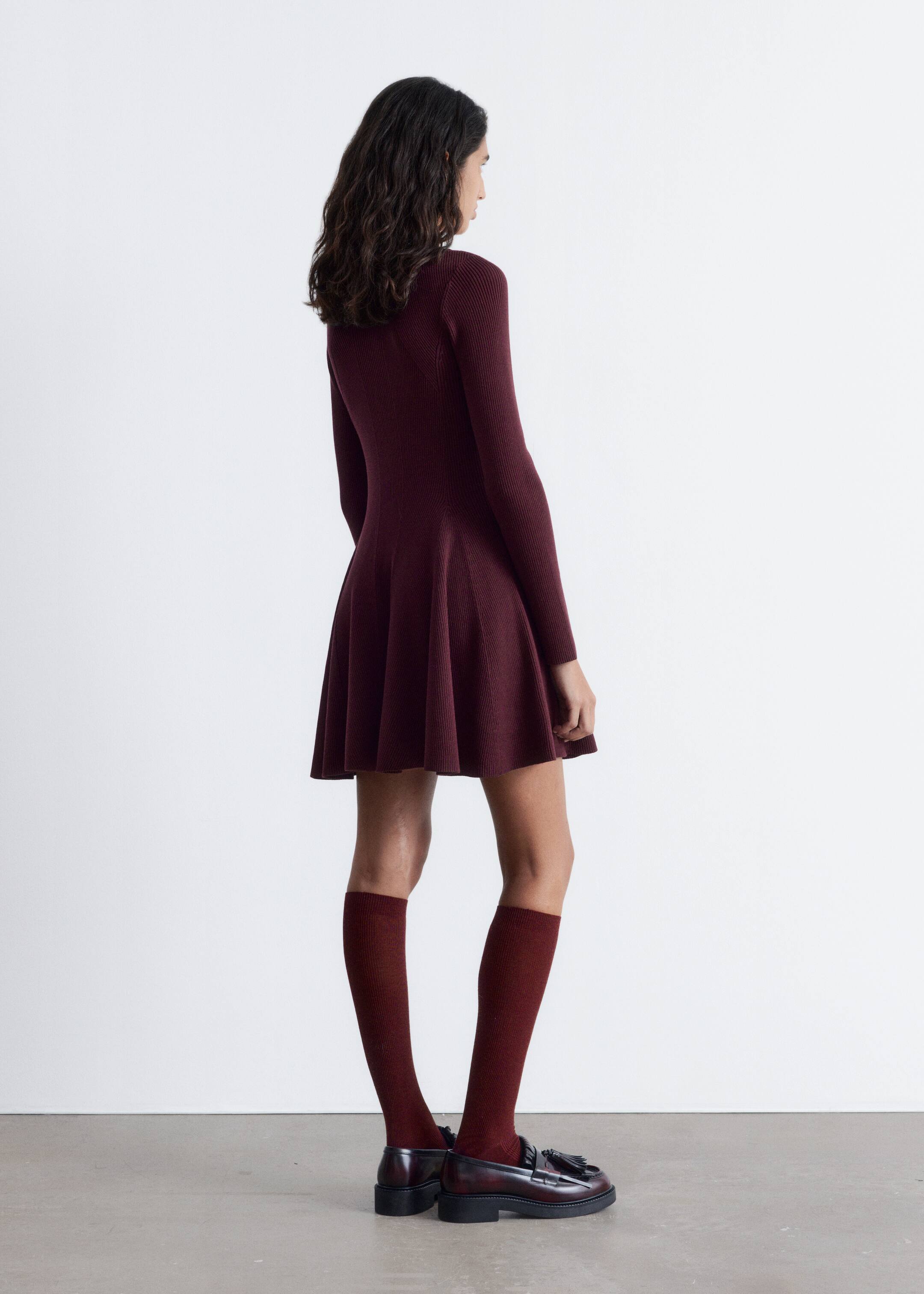 Agrandir l'image: Une robe mini à col montant en tricot côtelé rouge profond est portée par un mannequin. La robe présente une taille ajustée et une jupe évasée à panneaux, stylisée avec des chaussettes montantes assorties et des mocassins foncés.