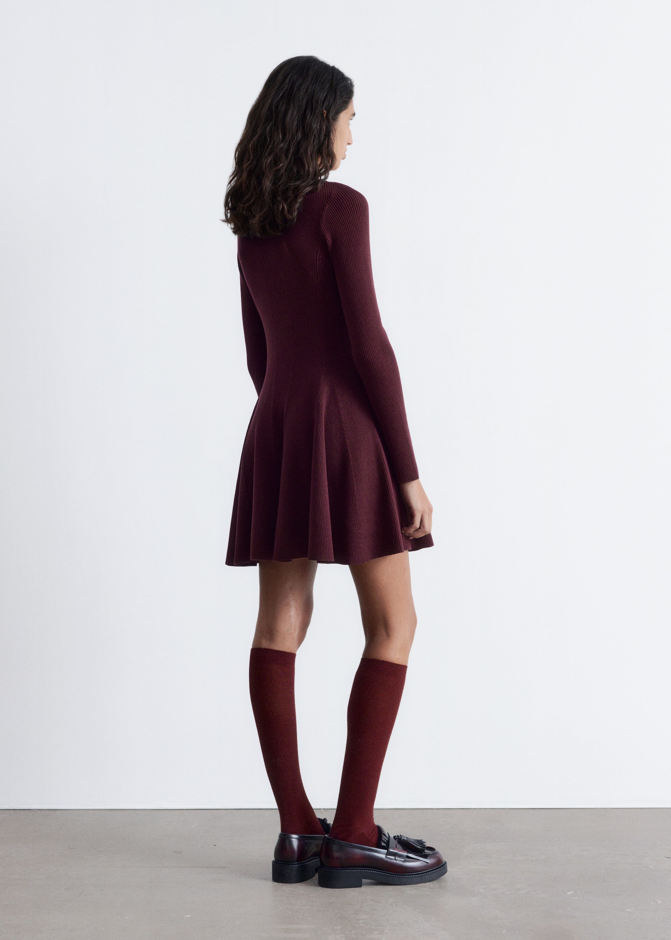 Se større billede: En dyb rød ribstrikket mock neck mini kjole bæres af en model. Kjolen har en tætsiddende talje og en udsvinget, paneleret nederdel, stylet med matchende knæhøje sokker og mørke loafers.