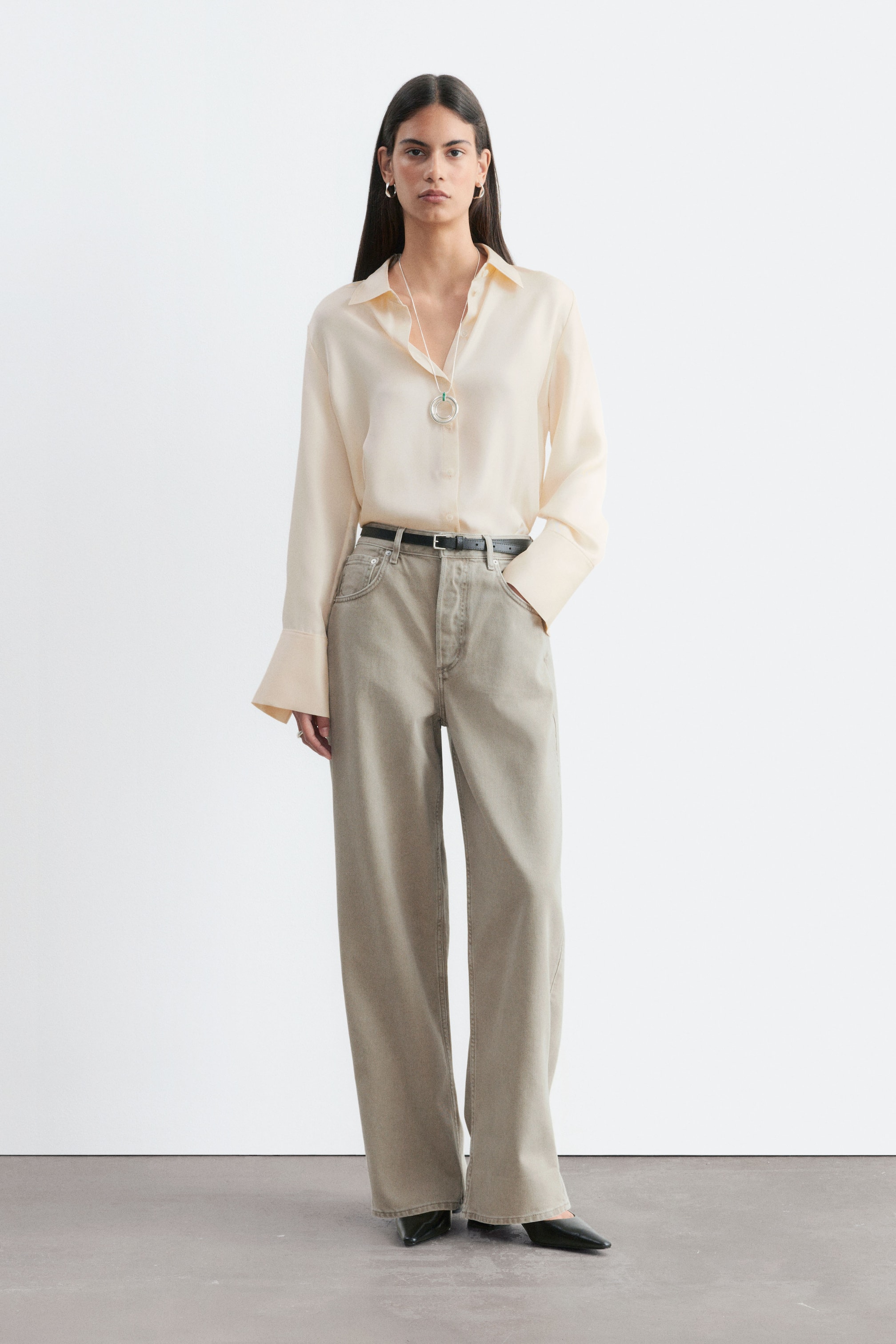 Größeres Bild ansehen: Die Model trägt eine hell khaki-graue Wide-Leg-Jeans, kombiniert mit einer in den Bund gesteckten cremefarbenen Langarmbluse und einem schmalen schwarzen Gürtel. Die Jeans zeichnen sich durch eine oversized, barrel-förmige Silhouette mit extra langen Beinen aus, die über schwarze Schuhe mit spitzer Zehenpartie fallen.
