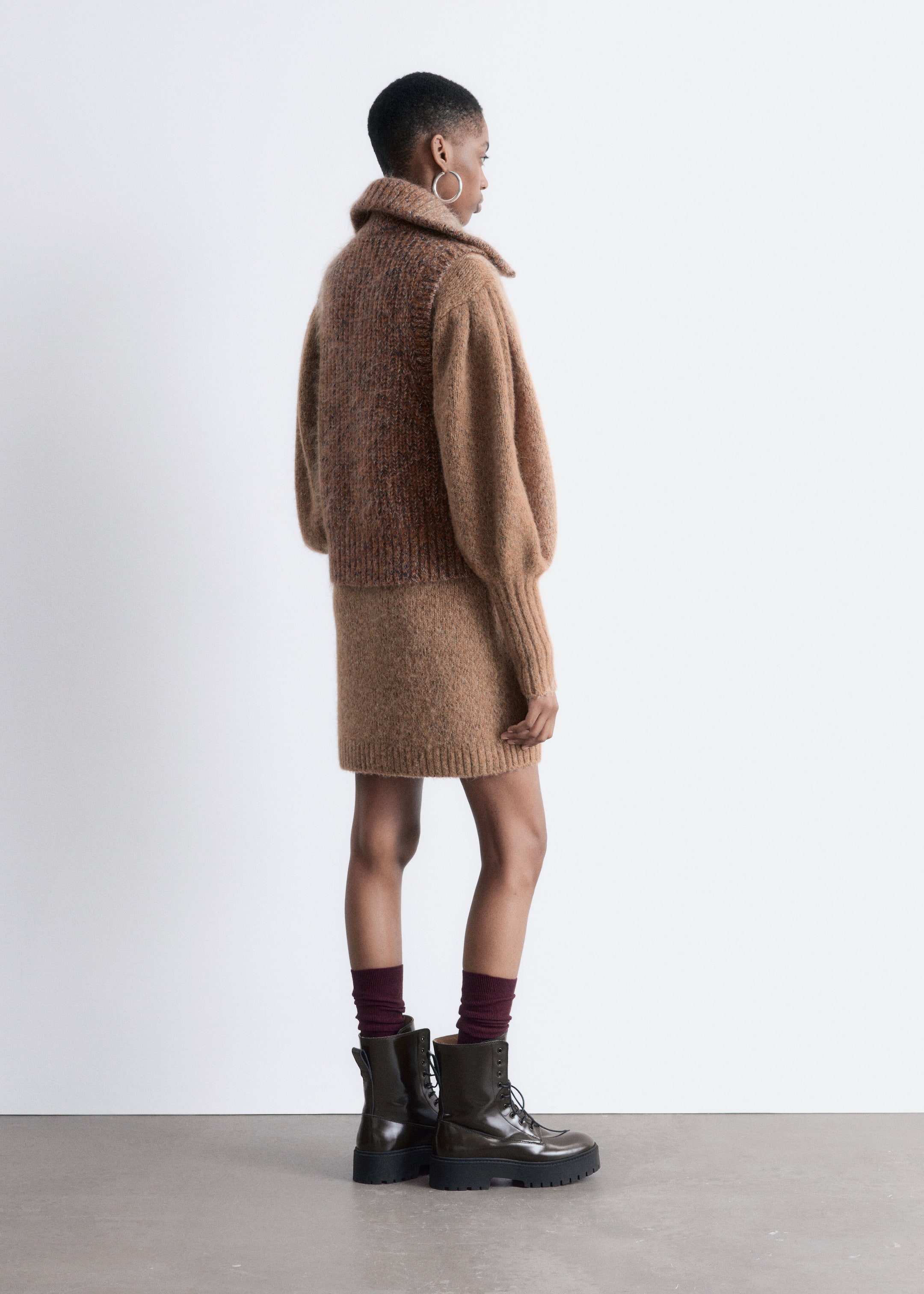 View larger image: A knitted sweater and skirt set is being shown from a side view. The short, chunky sweater is designed with a wide collar and voluminous long sleeves with ribbed cuffs. Its speckled brown knit body is contrasting with solid light brown sleeves. The coordinating short, straight-cut knit skirt in solid light brown is being styled with ribbed dark red socks and dark lace-up combat boots with thick lugged soles.