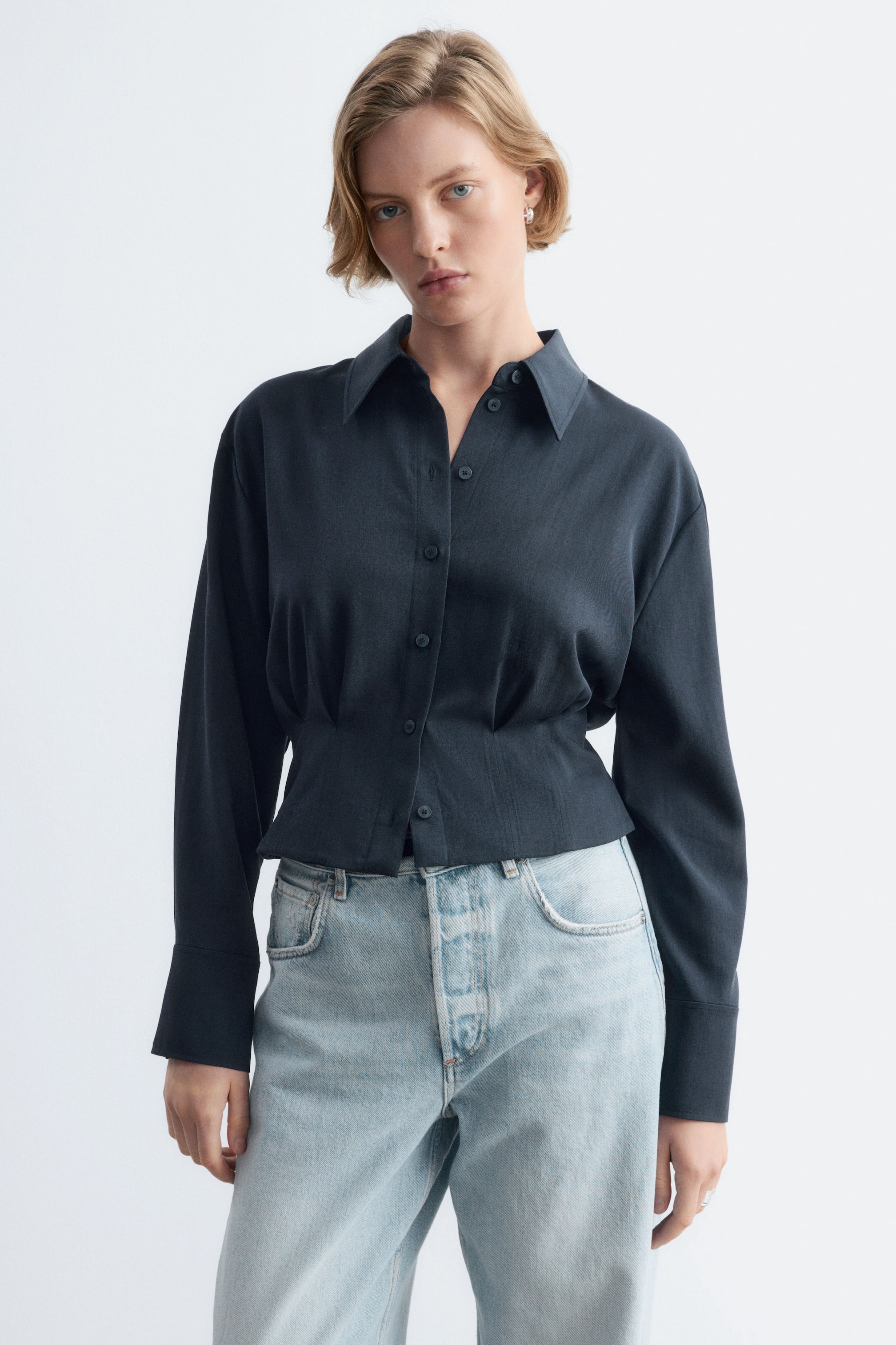 Agrandir l'image: Une chemise à manches longues gris foncé, dotée d’un col net, d’une patte de boutonnage sur le devant et de plis à la taille qui structurent sa silhouette, est portée par le mannequin. La chemise est associée à un jean en denim délavé clair.