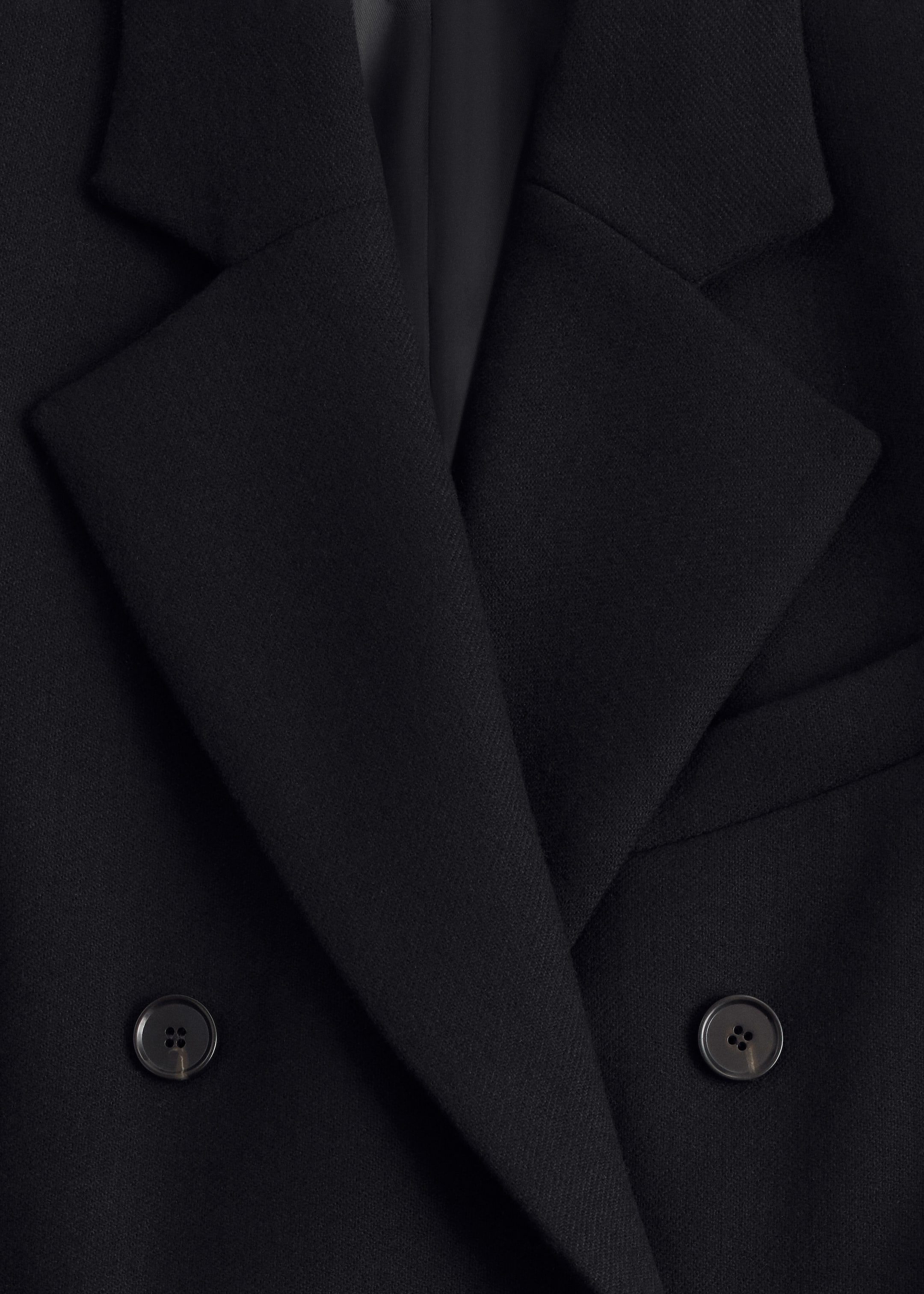View larger image: A close-up view is showing the upper portion of a black double-breasted coat, featuring wide notch lapels and two dark, four-hole buttons on its overlapping front panels.