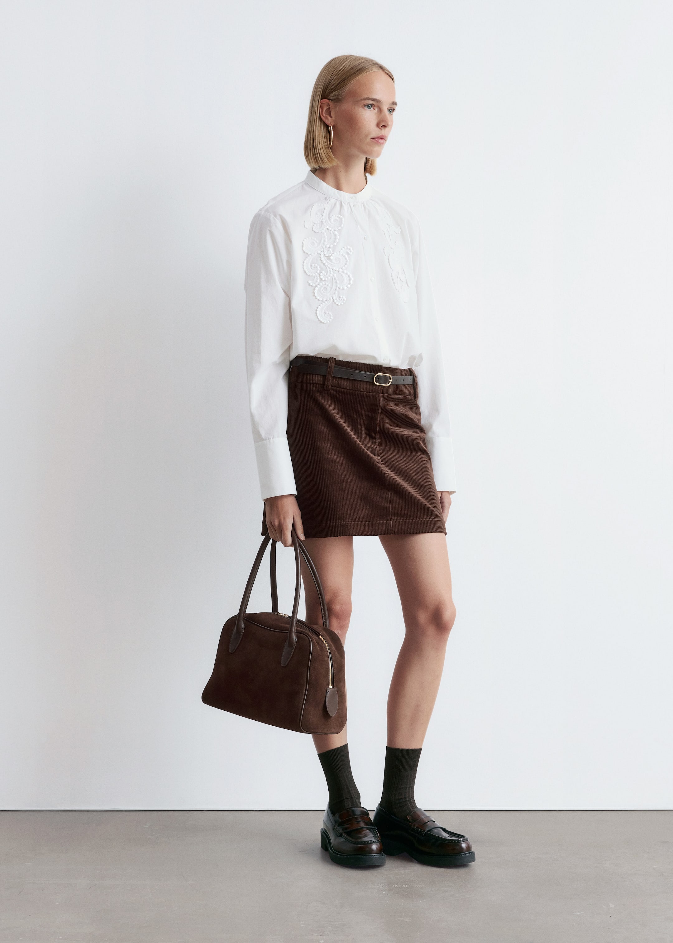 View larger image: A white long-sleeved shirt with a band collar and white floral embroidery on the chest is being worn tucked into a brown corduroy mini skirt with a slim dark belt. The ensemble is completed with dark ribbed socks, chunky-soled dark brown penny loafers, and a dark brown suede tote bag with leather handles being held.
