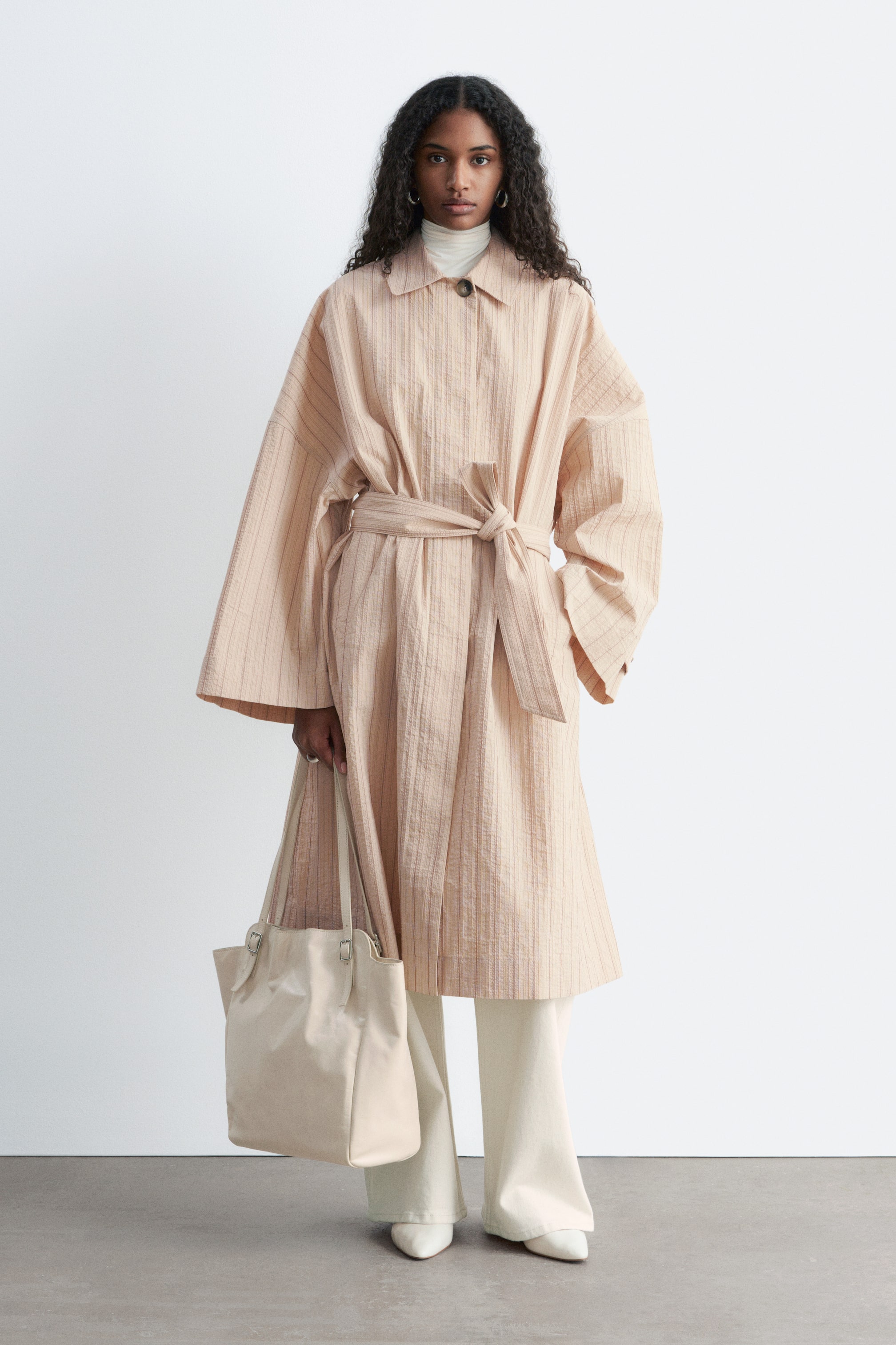 Se større billede: Den ferskenfarvede frakke har en subtil lodret nålestribet tekstur, en spids krave og brede ærmer, der vender fremad. Den er designet med et selvbindende bælte, der strammer taljen ind, og en enkelt synlig knaplukning øverst, lagdelt over en hvid rullekrave.