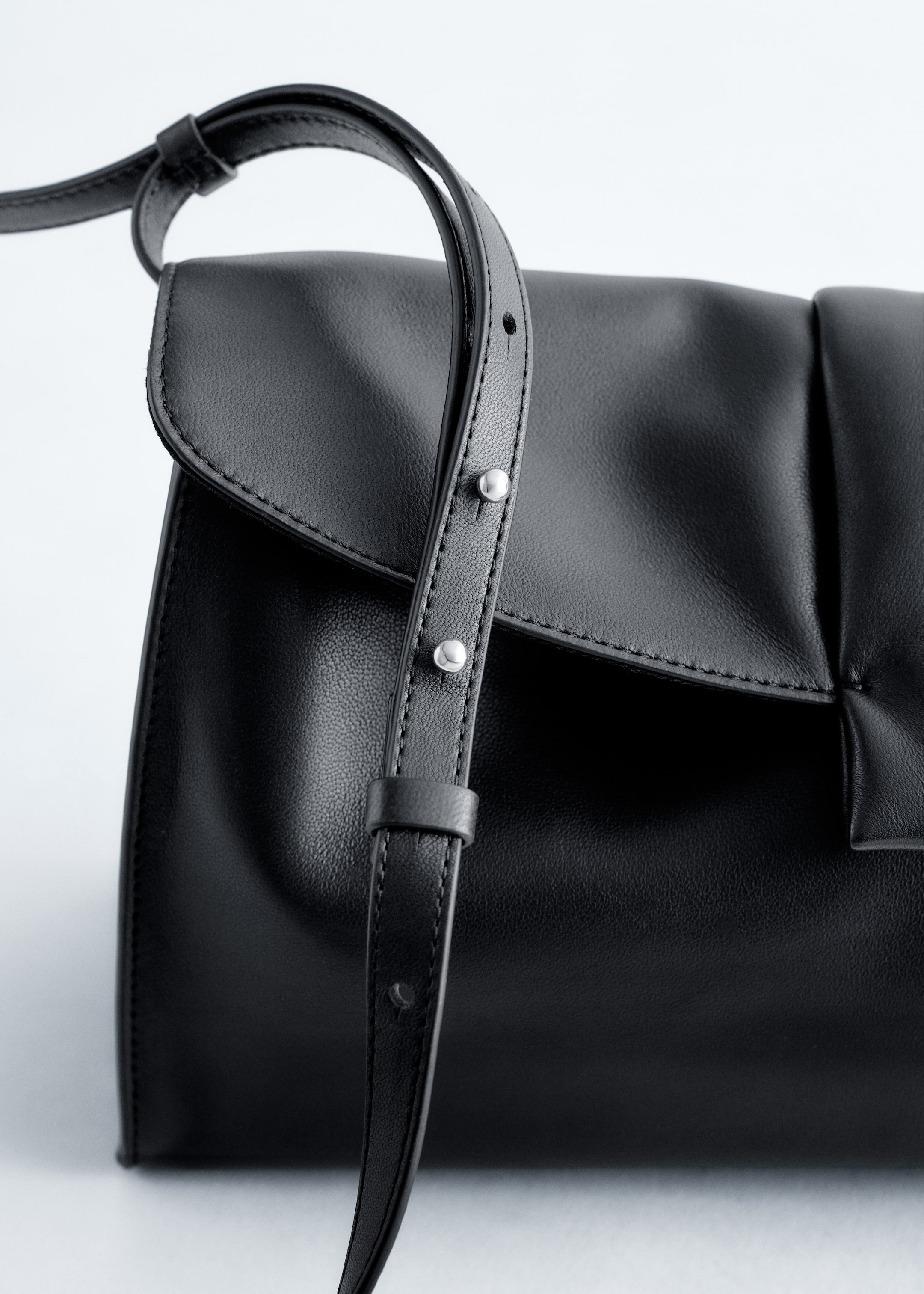Größeres Bild ansehen: Eine schwarze Lederclutch wird aus einem schrägen Blickwinkel gezeigt, mit einer weichen, geschmeidigen Textur und einem gebogenen Klappenverschluss. Ein verstellbarer Schulterriemen mit silberfarbenen Nieten-Details ist über die Vorderseite der Tasche drapiert.