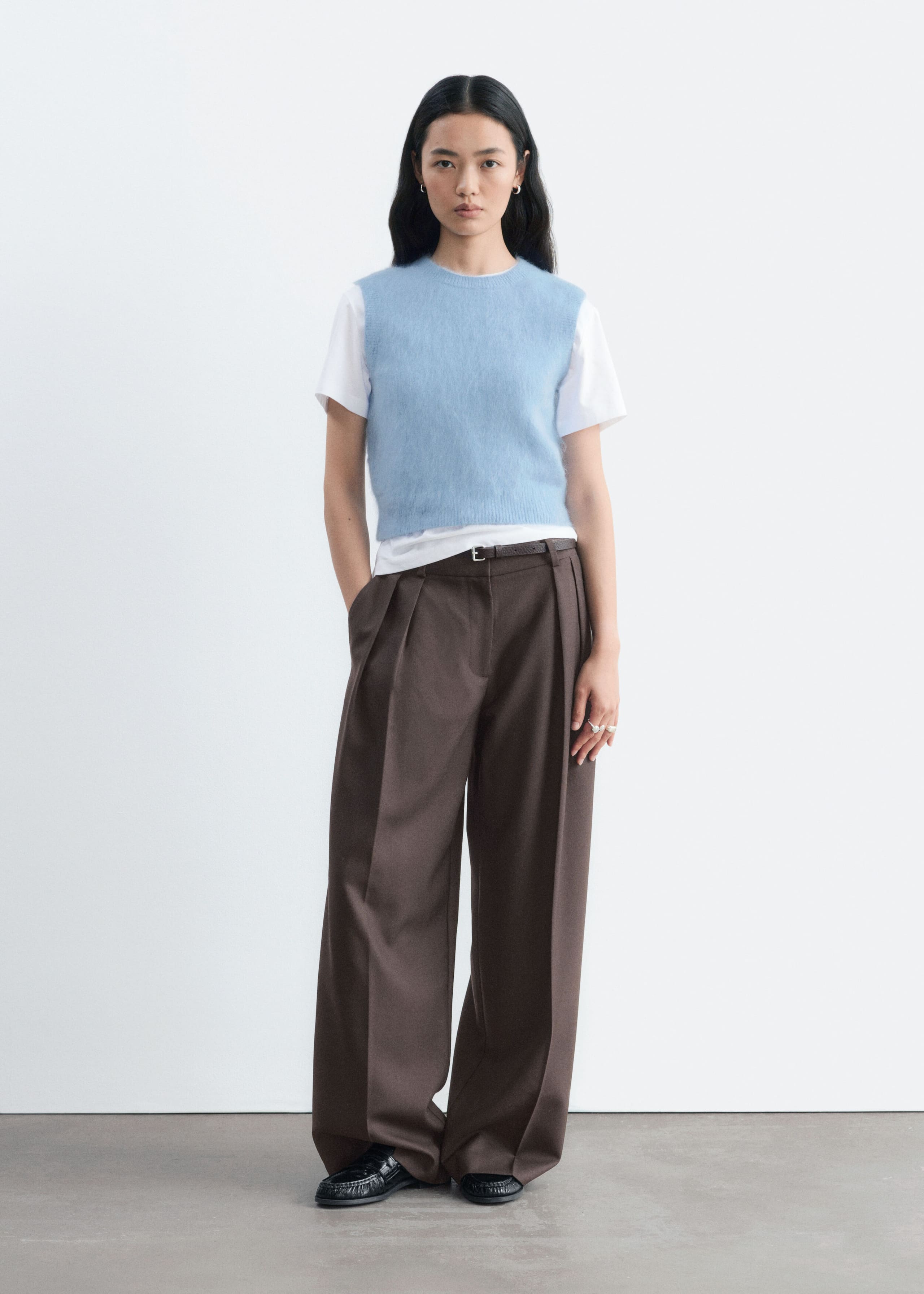 La modelo lleva un chaleco de mezcla de mohair de color azul claro, con cuello redondo, diseño sin mangas y un acabado suave con textura. El chaleco cropped se superpone sobre una camiseta blanca de manga corta y se combina con unos pantalones marrones anchos de pinzas.