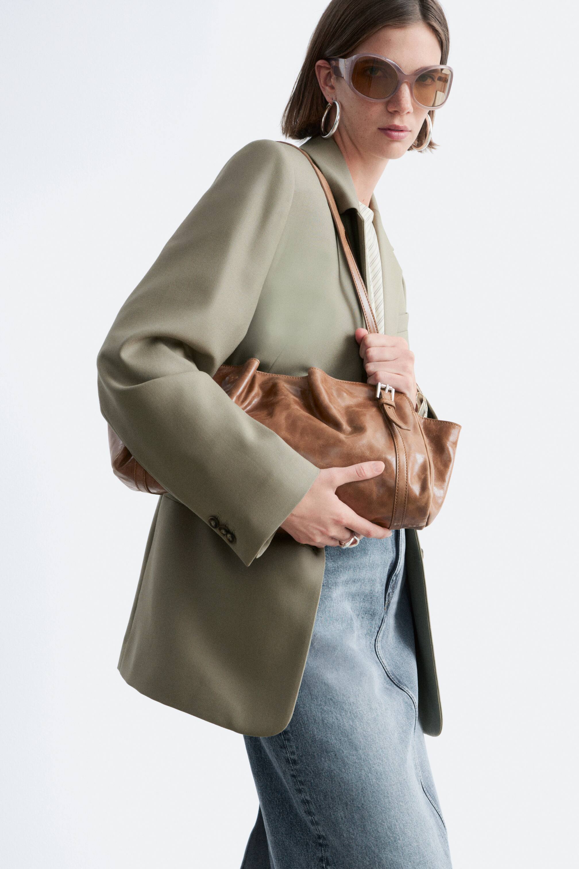 Visa större bild: En brun axelväska i läder, med en mjuk, rynkad silhuett och en justerbar rem med ett spänne i silverton, bärs och hålls av modellen.