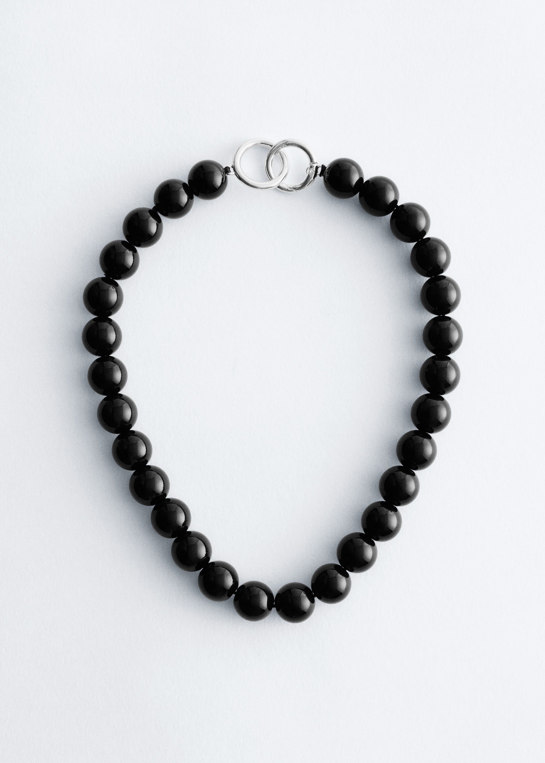 Visualizza immagine più grande: Una collana presenta numerose perle nere lucide e tonde. Un fermaglio a O di colore argento collega le estremità del filo di perle, che giace piatta in una forma circolare aperta.