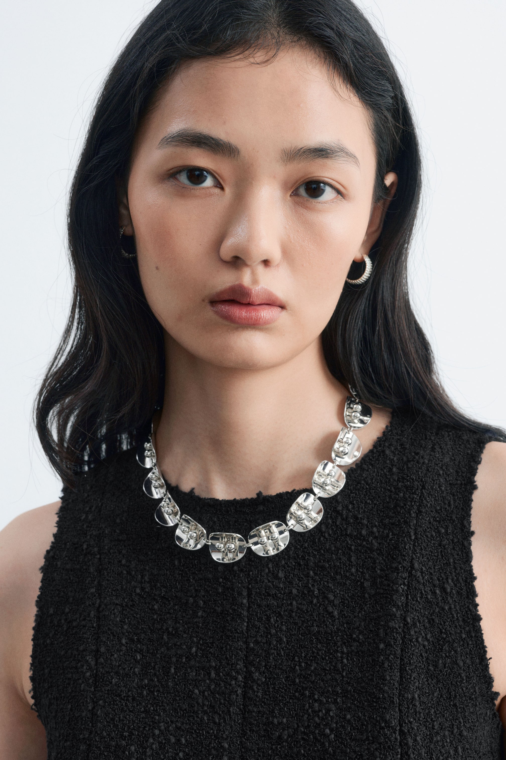 Agrandir l'image: Un collier ton argenté, à l’aspect poli, composé de panneaux cintrés reliés entre eux et agrémenté de détails sphériques, est porté par le mannequin. Le collier est associé à une mini-robe noire sans manches à la texture travaillée, avec un col à bord brut.