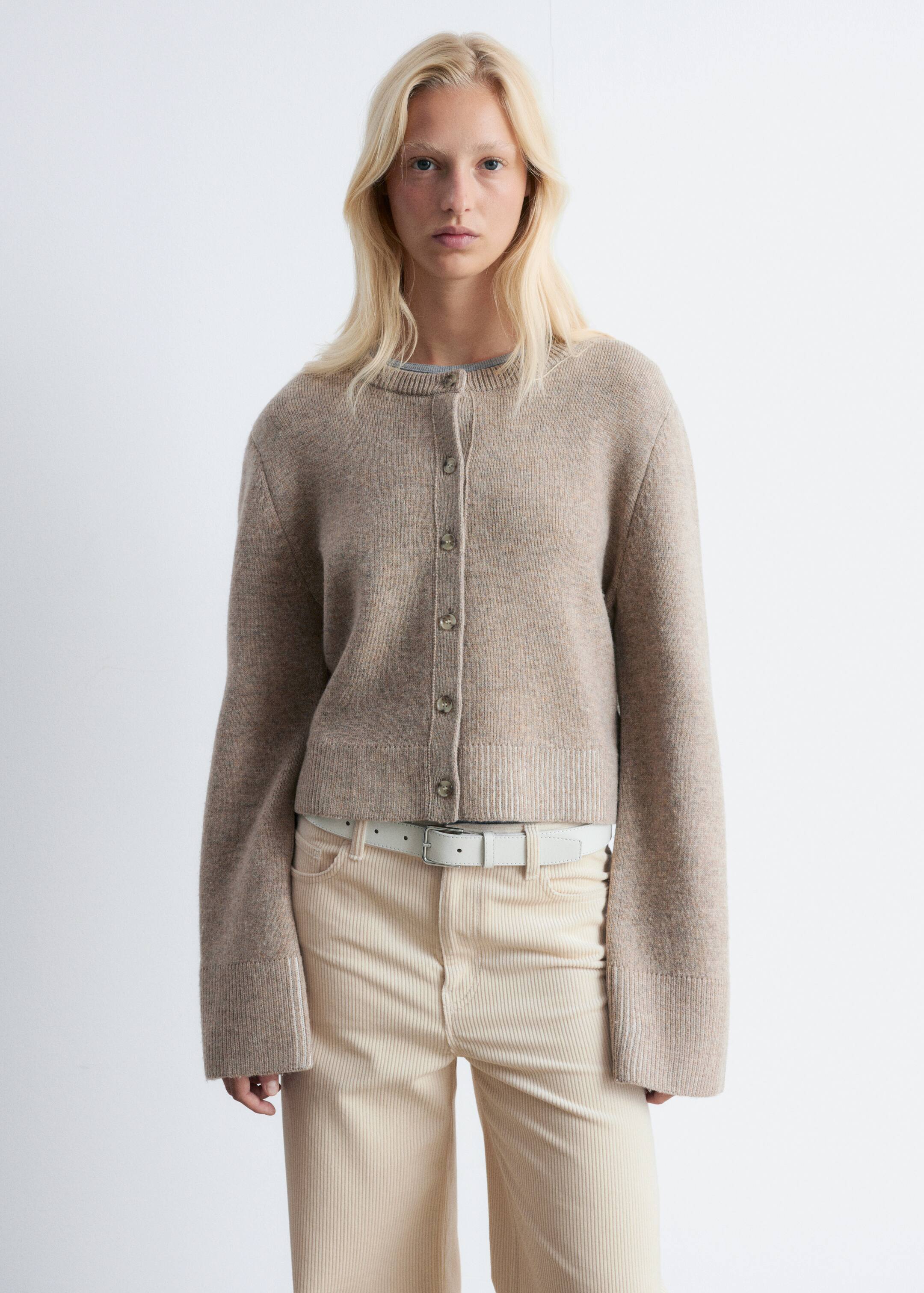 View larger image: A beige heathered knit cardigan is being shown front-facing, featuring a crew neck and a button-front placket with light-colored buttons. It is designed with wide, long sleeves that have distinct hems and a ribbed bottom hem, appearing slightly cropped and layered over a lighter top visible at the neckline.