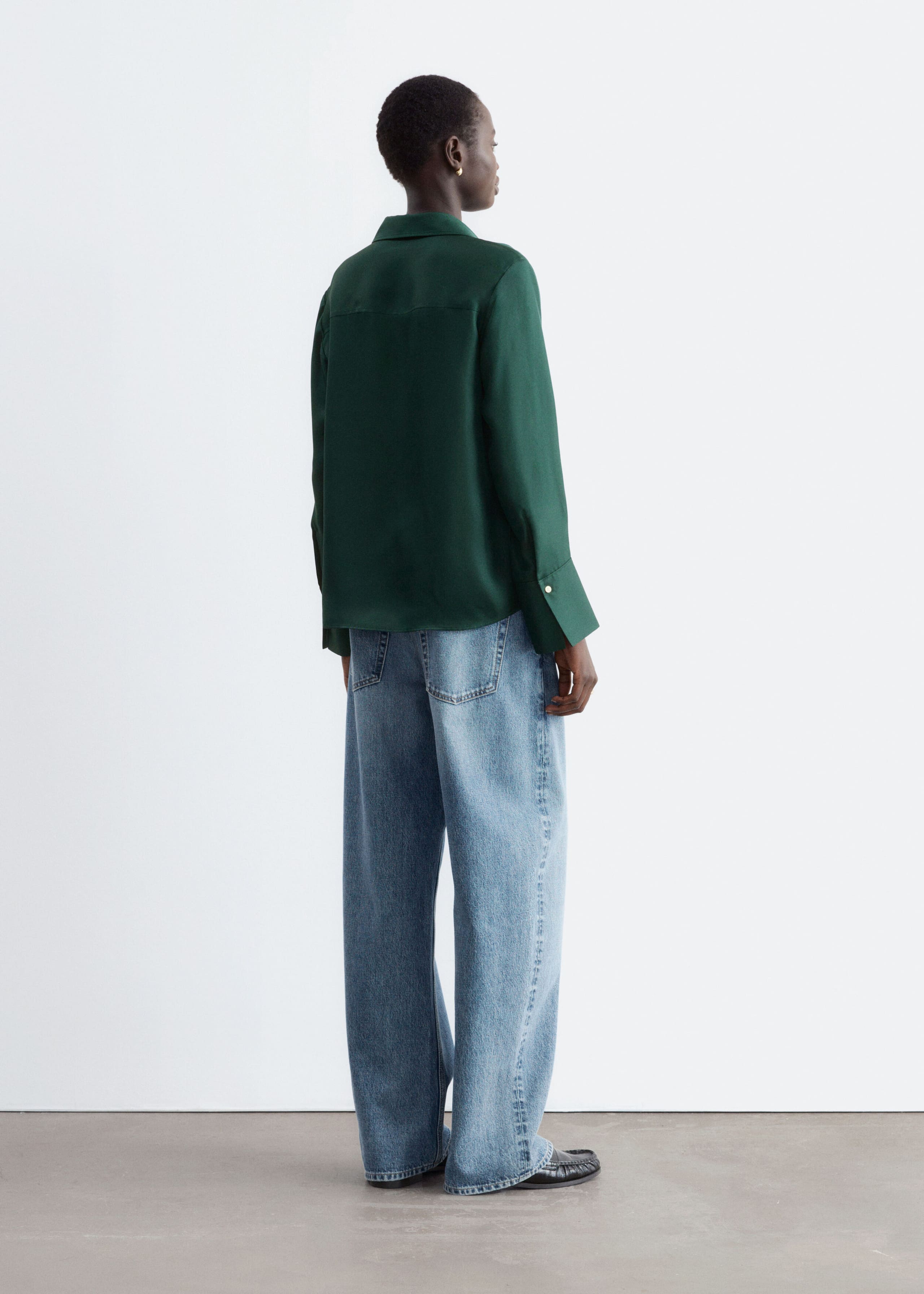 A dark green silk shirt featuring a relaxed silhouette, a back yoke, and wide cuffs is worn by the model. The shirt is paired with wide-leg blue jeans.